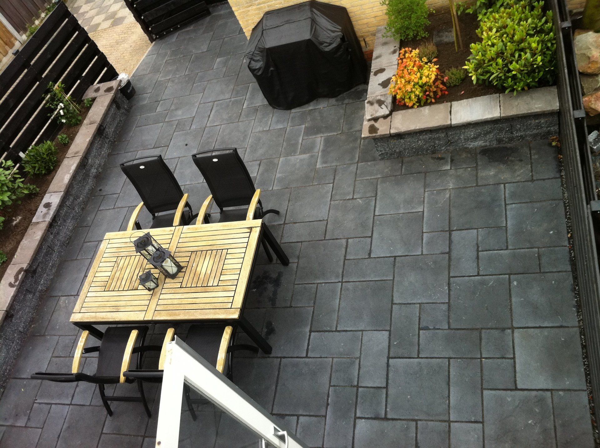 Terras met donkergrijze bestrating, een houten tafel, stoelen en een bloembed met groen.