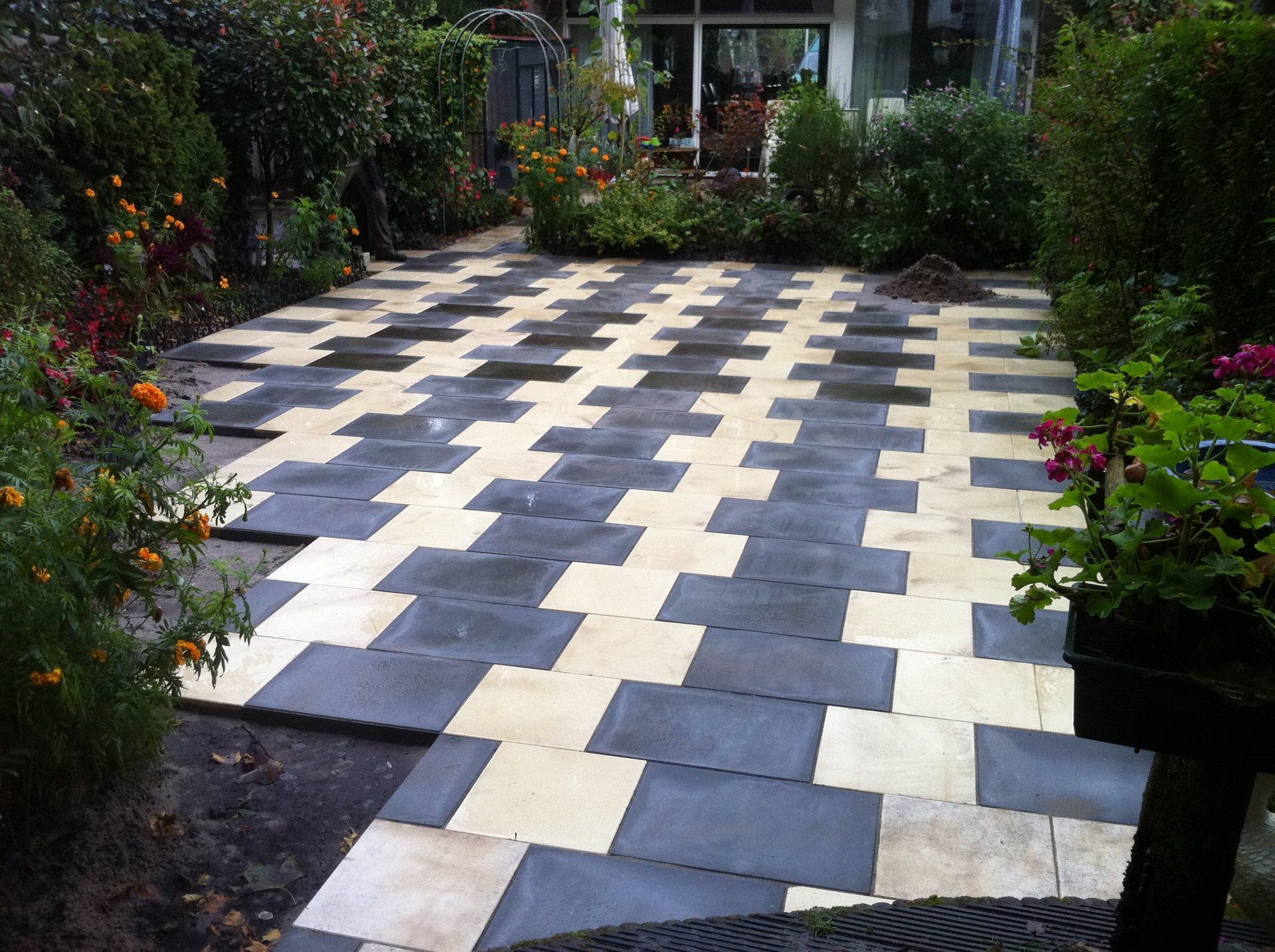 Terras bestraat met afwisselend lichte en donkere rechthoekige blokken, omgeven door planten en struiken.