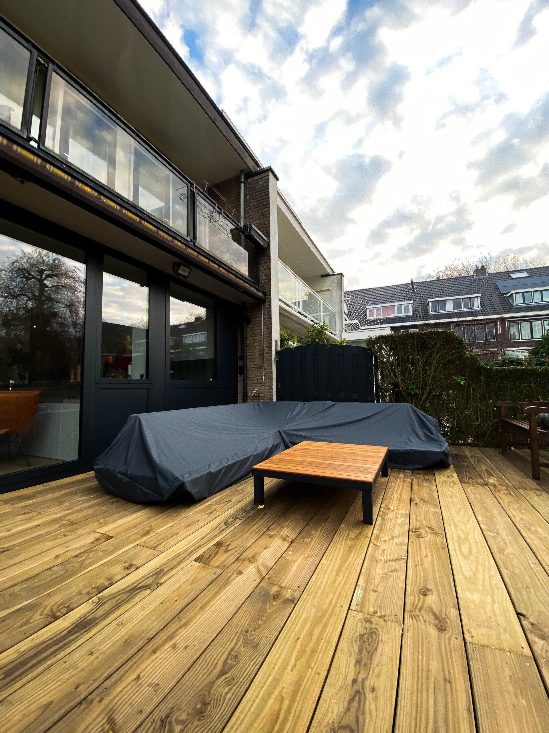 Houten terras met overdekt tuinmeubilair en een klein houten tafeltje, naast een modern gebouw.