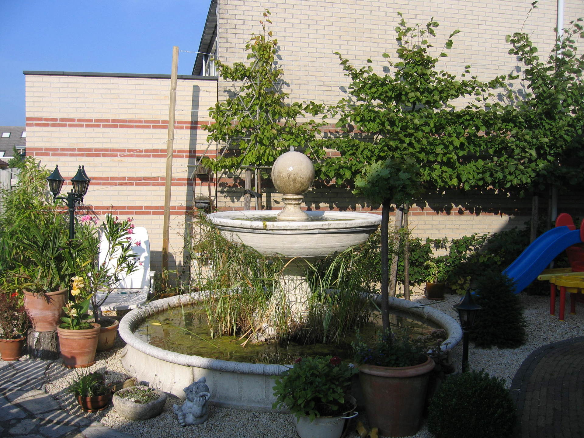 Fontein in een tuin met bomen, potplanten en een bakstenen muur op de achtergrond.