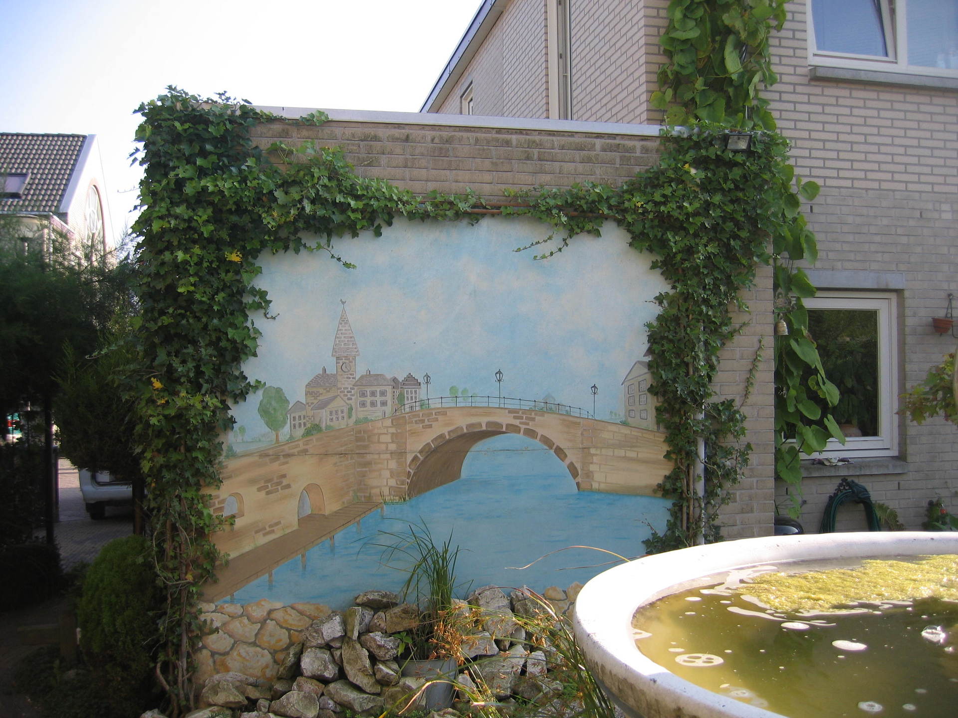Muurschildering van een brug over een rivier, omlijst door groene klimplanten, naast een bakstenen gebouw.