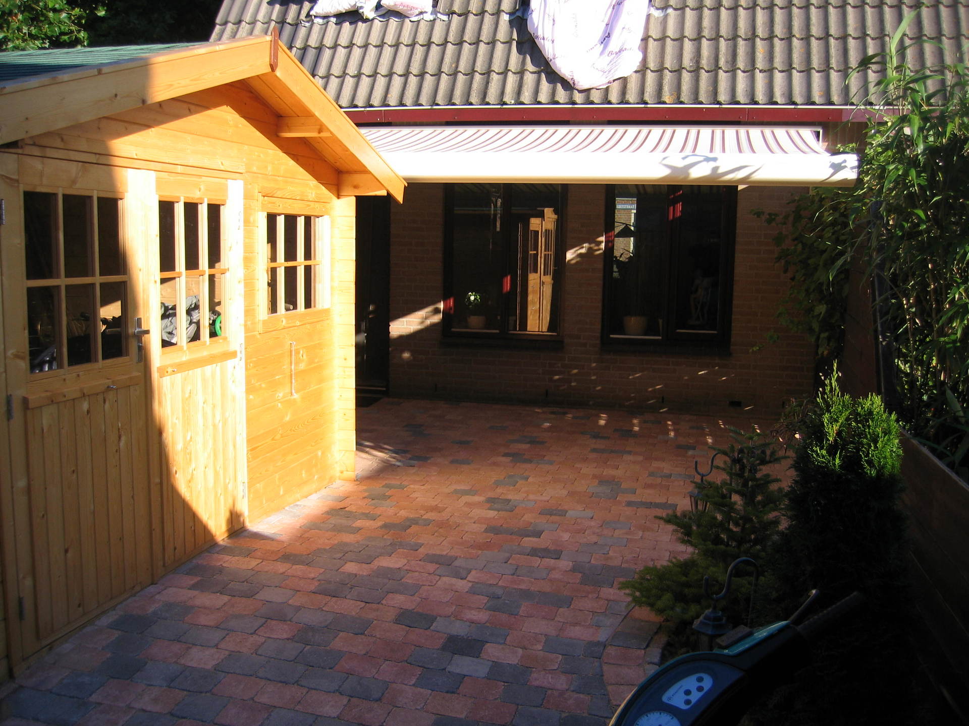 Houten schuur en stenen terras in een zonnige achtertuin met een gebouw en luifel op de achtergrond.