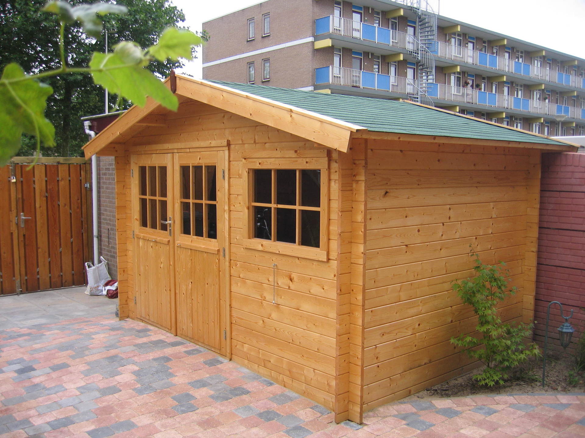 Houten schuur met dubbele deuren en raam, groen dak, gelegen op een stenen terras met een gebouw op de achtergrond.