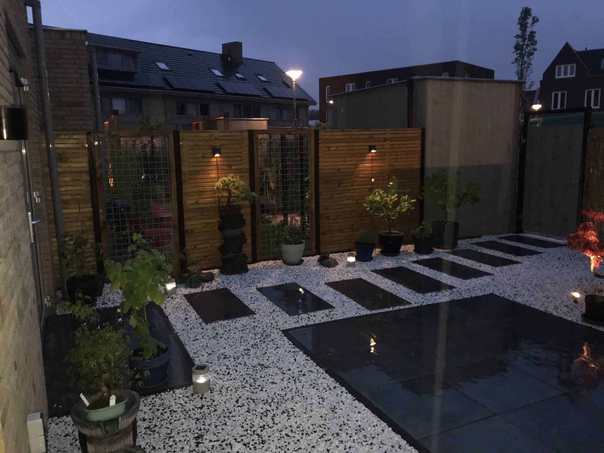 Een moderne achtertuin in de schemering met houten schuttingen, donkere tegels, witte stenen en sfeerverlichting.