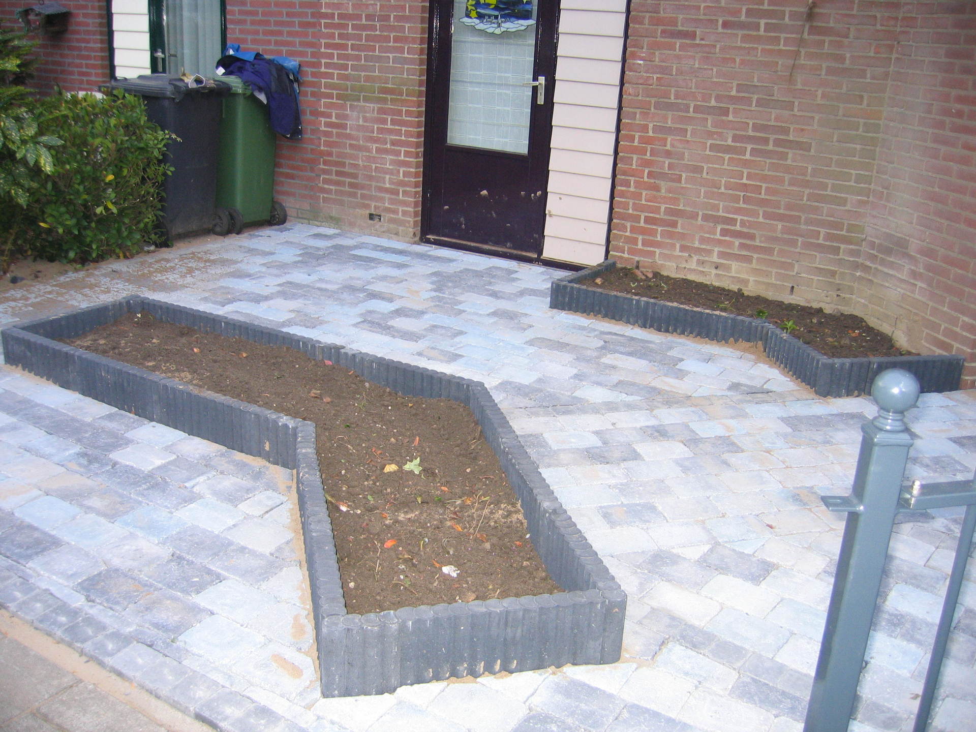 Een stenen terras met twee hoekige, grijze plantenbakken gevuld met aarde; op de achtergrond een stenen gebouw.