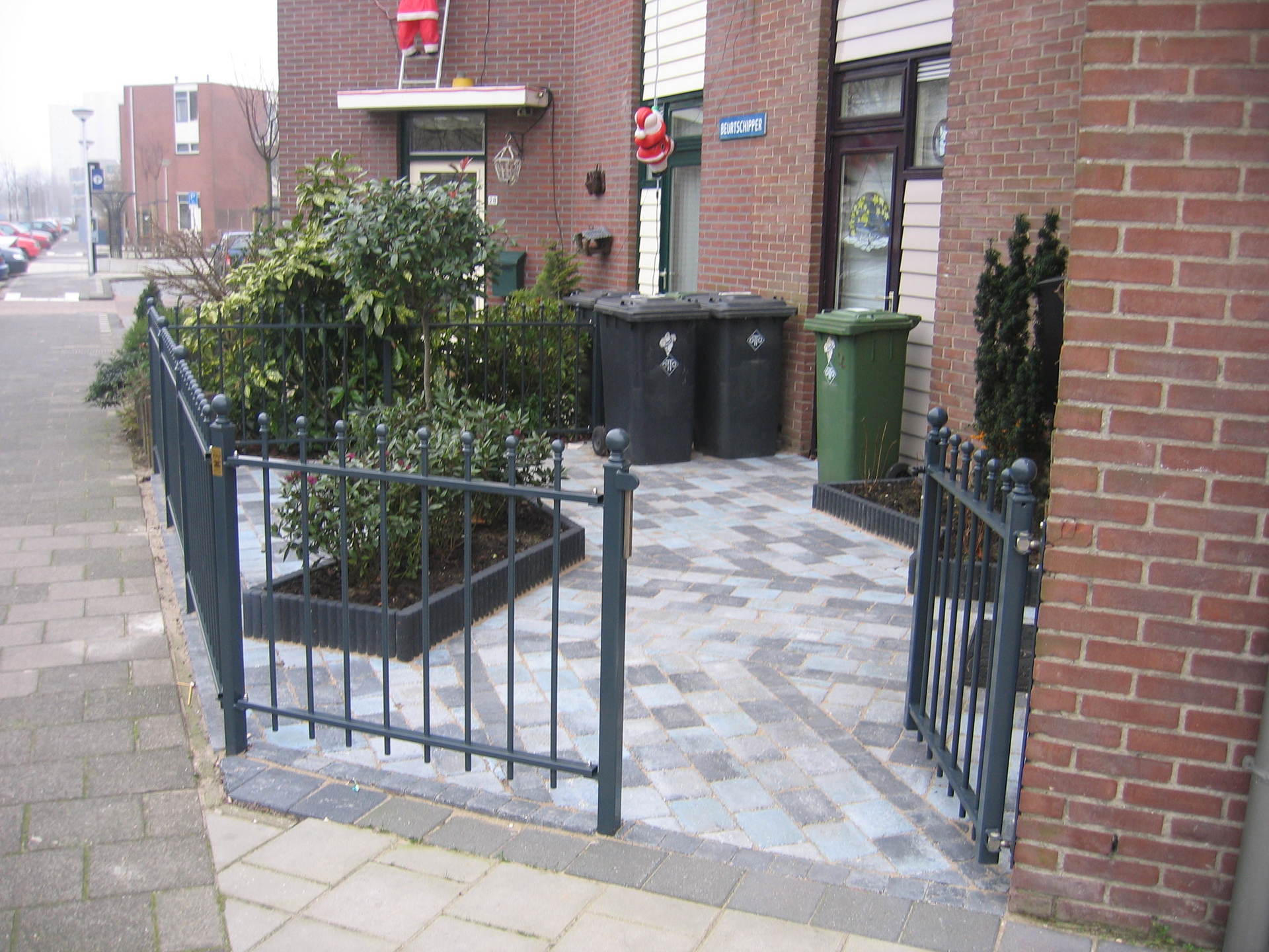 Ingang met bakstenen pad, zwart hek en afvalbakken bij een huis.