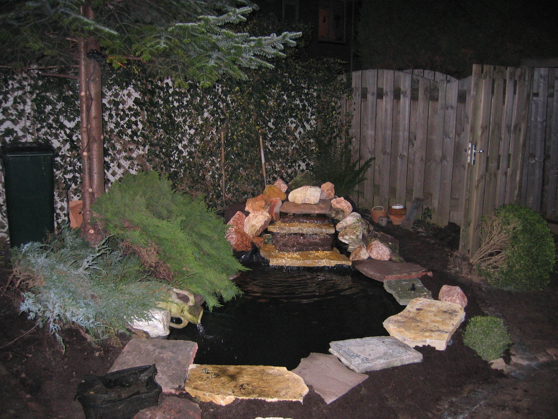 Een vijver in de achtertuin met een kleine waterval, omgeven door rotsen en planten, 's nachts verlicht door gedempt licht.
