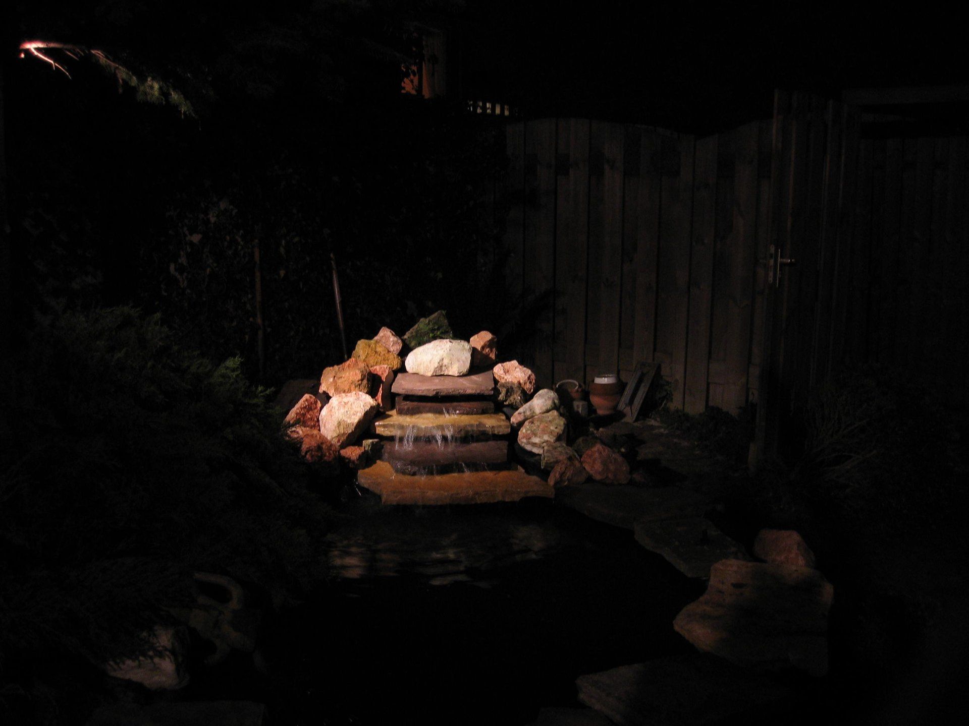 Een kleine, verlichte waterval in een vijver in een achtertuin, 's nachts. Donkere omgeving.
