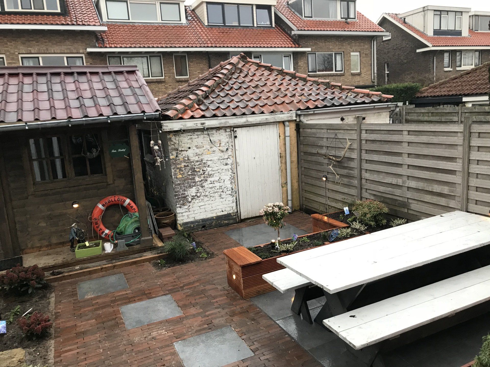 Achtertuin met stenen terras, verweerde schuurtjes, picknicktafel en schutting. Op de achtergrond zijn rode pannendaken te zien.