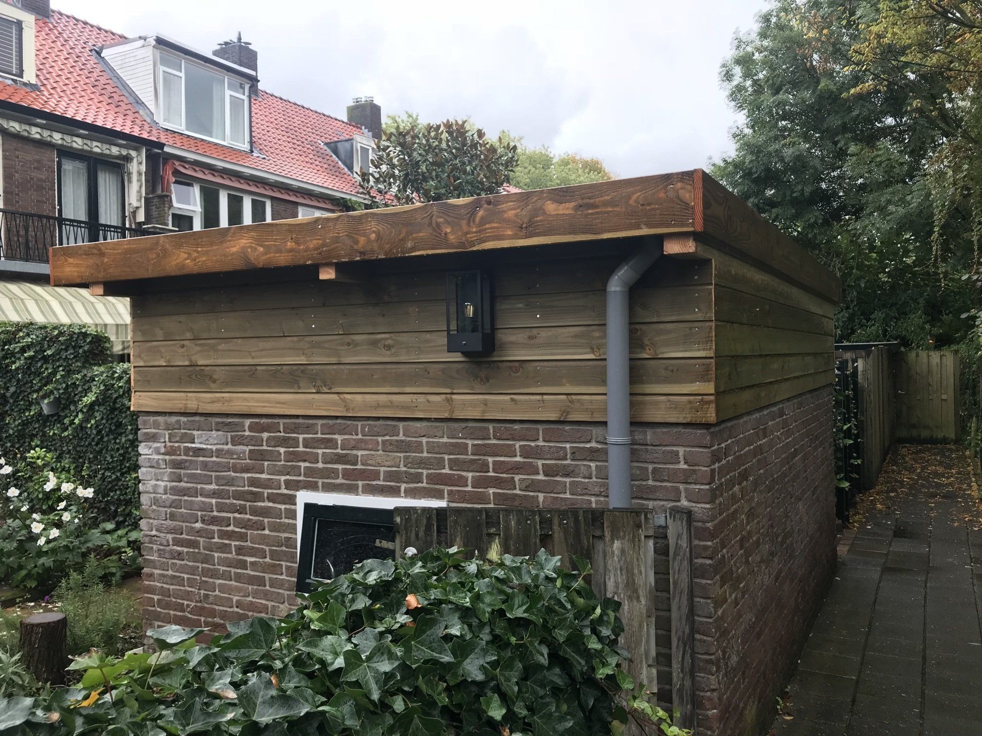 Een schuur van baksteen en hout met een plat dak, een grijze dakgoot, een klein raam en een deur.
