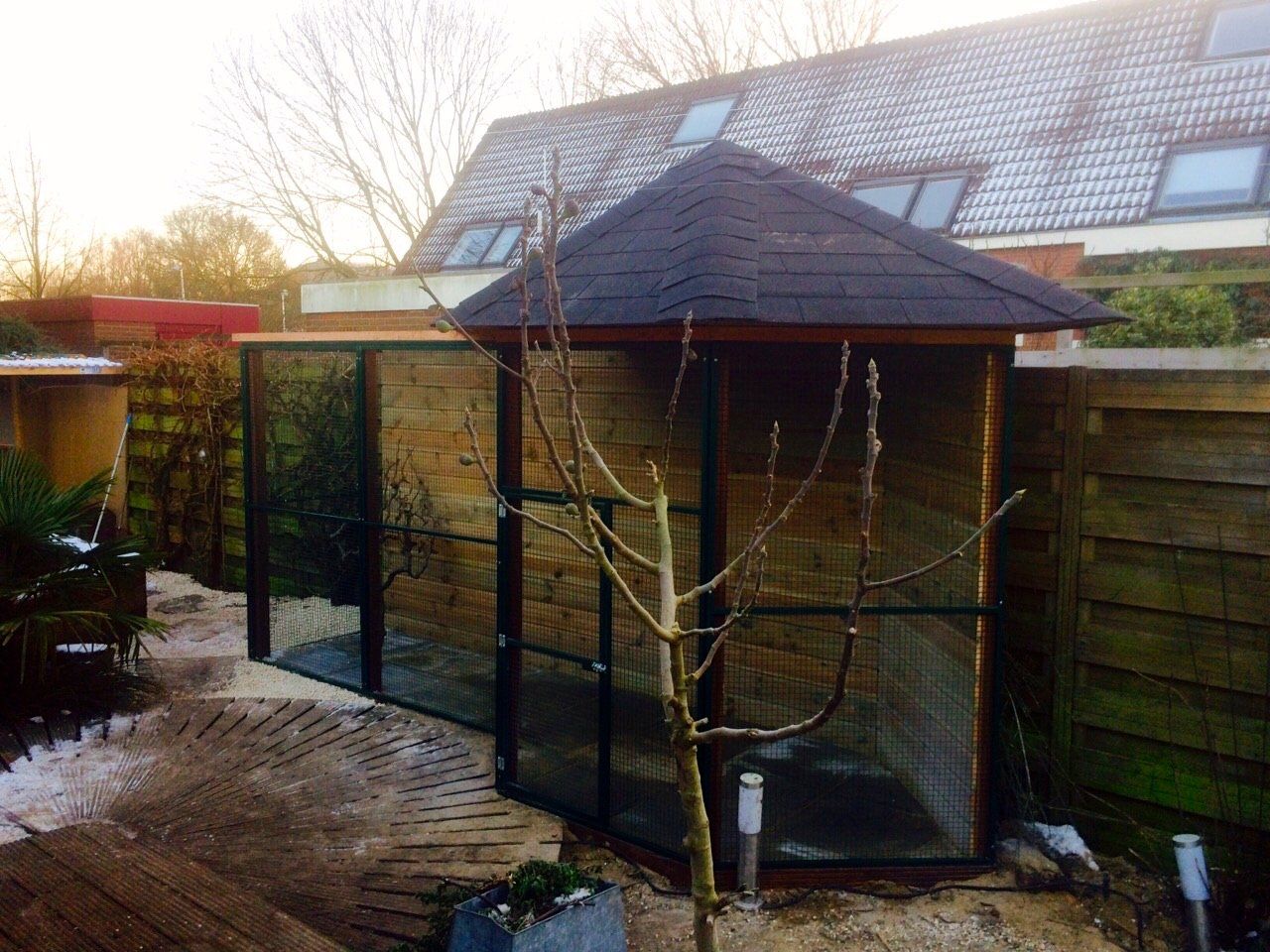 Een houten prieel en een afgesloten ruimte in een achtertuin, omgeven door groen en een bakstenen pad, met een besneeuwd dak op de achtergrond.