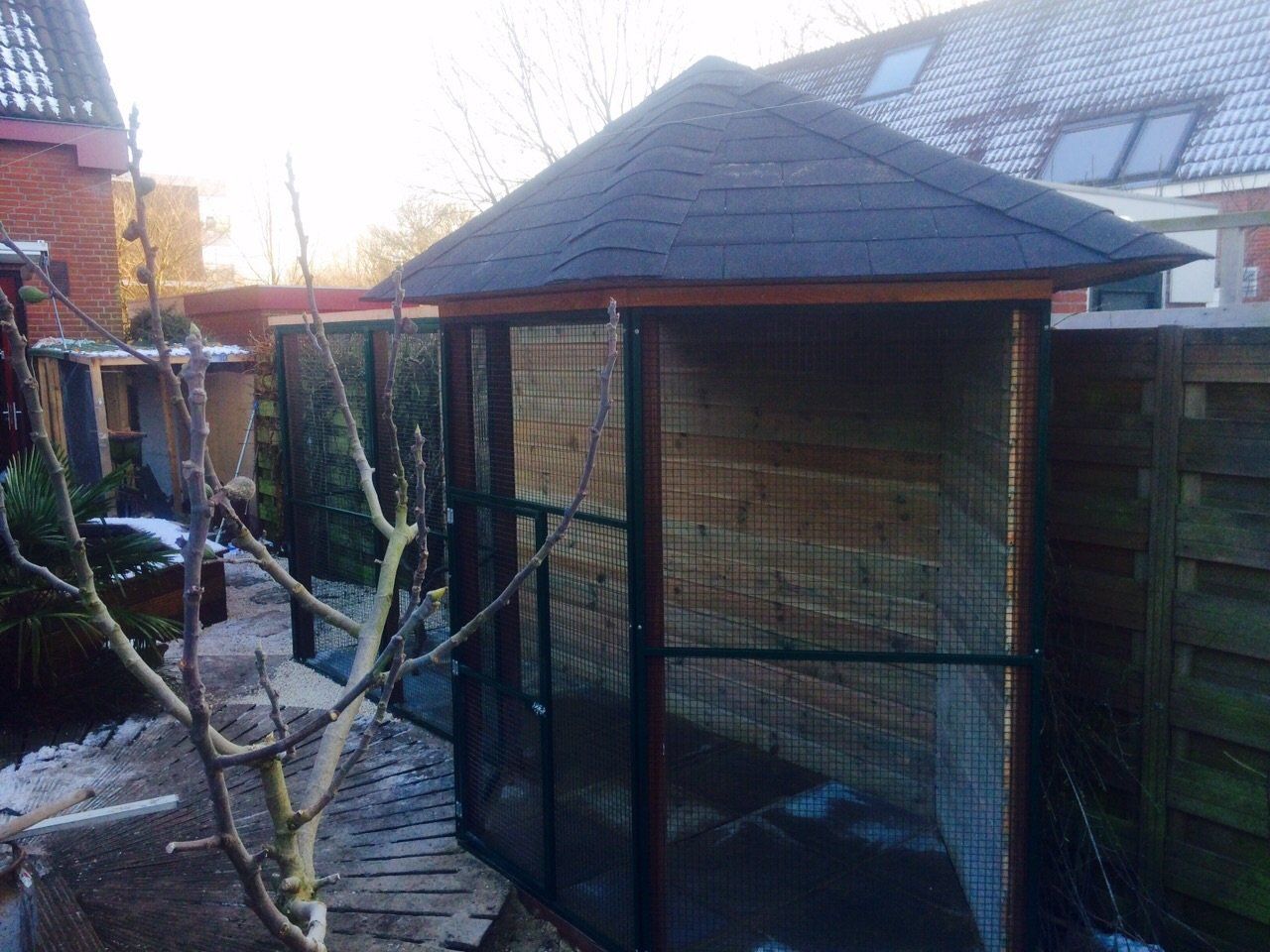 Een tuinpaviljoenachtige volière met gaaswanden en een donker dak in een achtertuin, vlakbij een houten schutting.