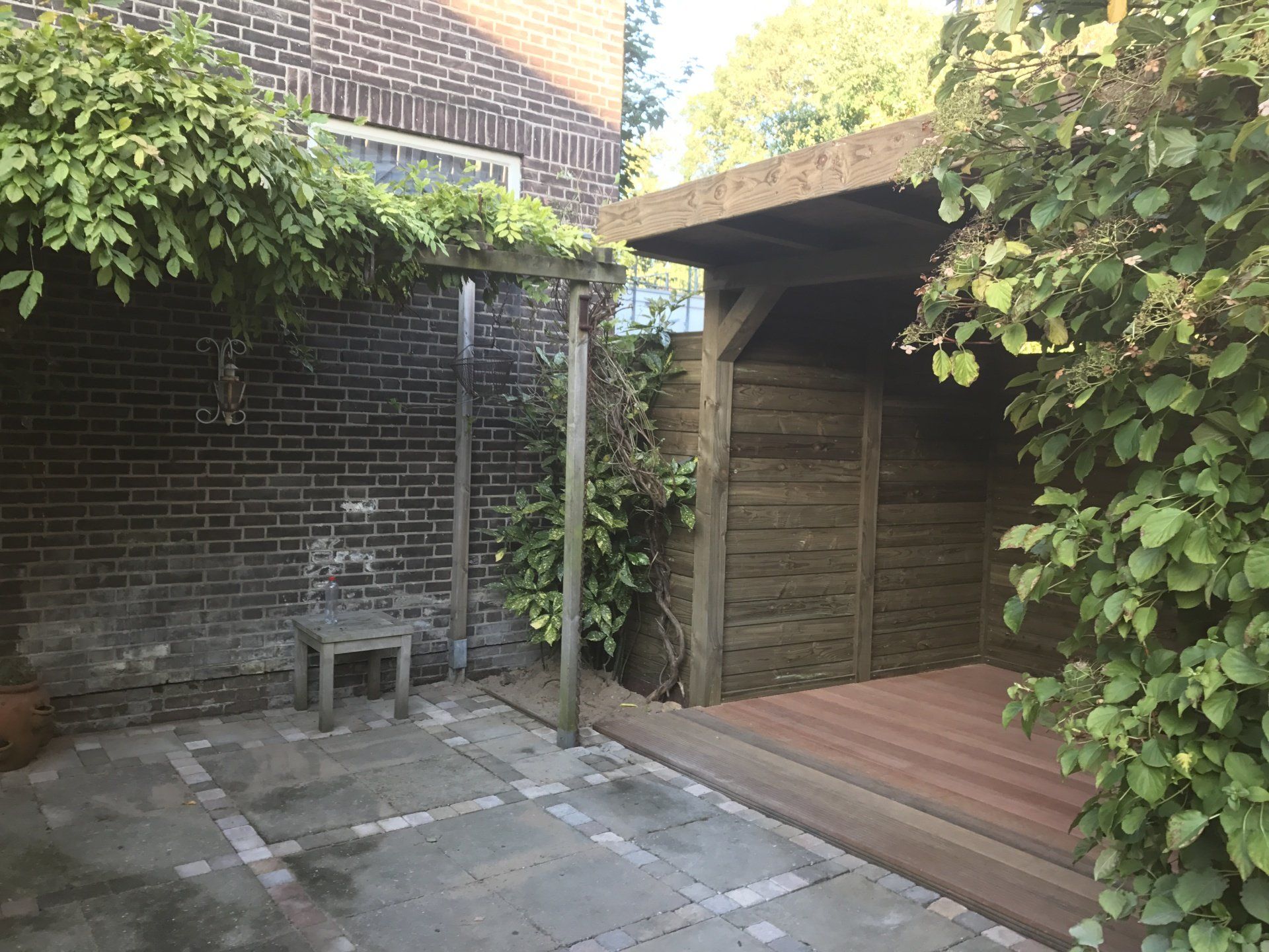 Terras met bakstenen muur, houten schuur, klimplanten en een kleine stoel.