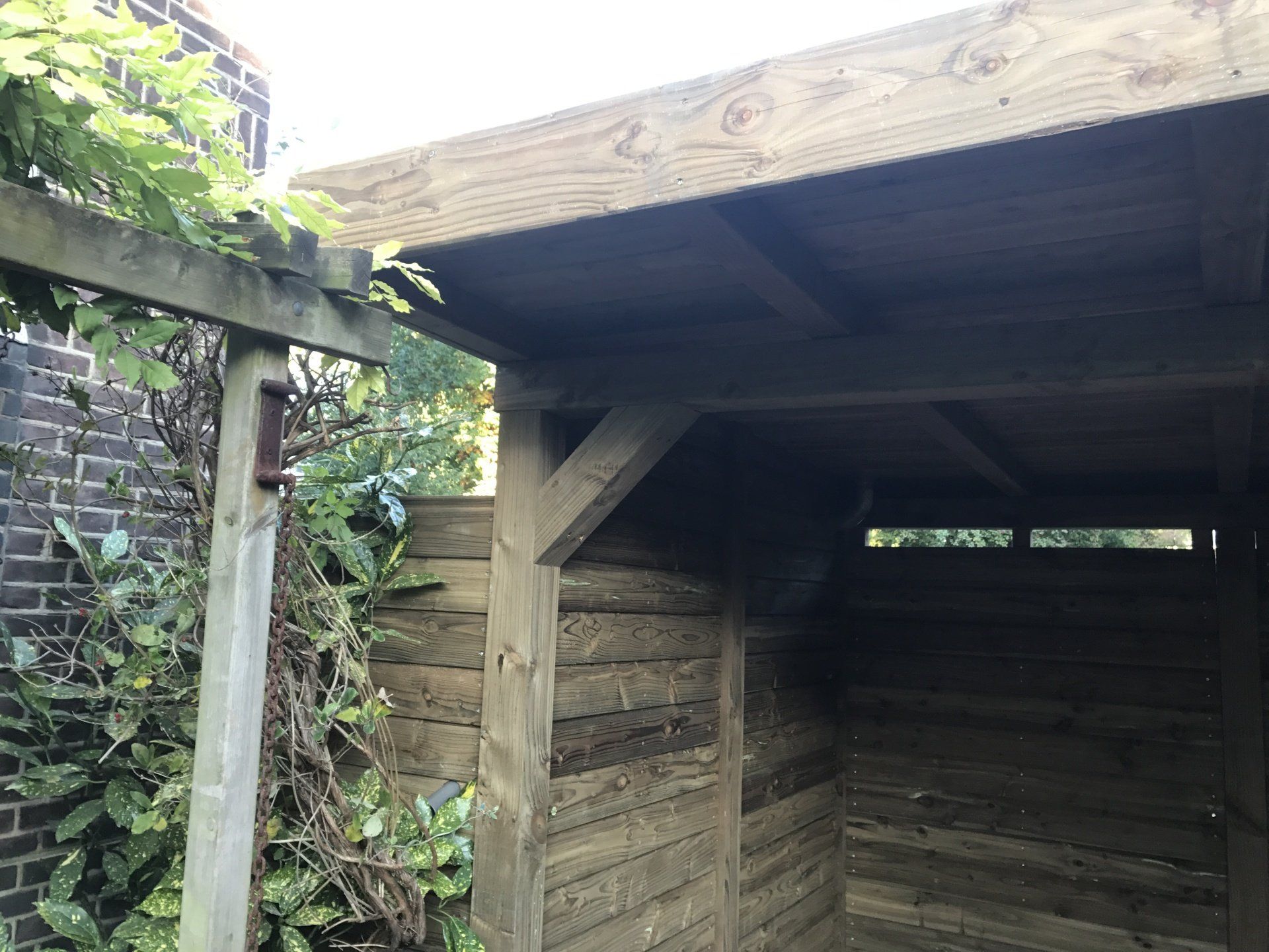 Houten schuur met een schuin dak in een tuin, met planten en een bakstenen muur.