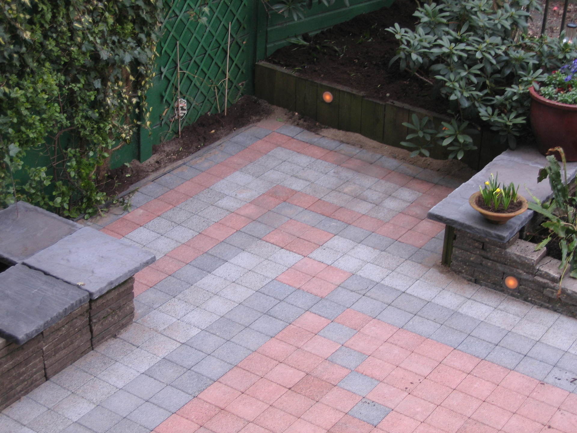 Een terras van bakstenen met een patroon van grijze en roodroze bakstenen, grenzend aan een bloembed met planten en een ingebouwde trede.