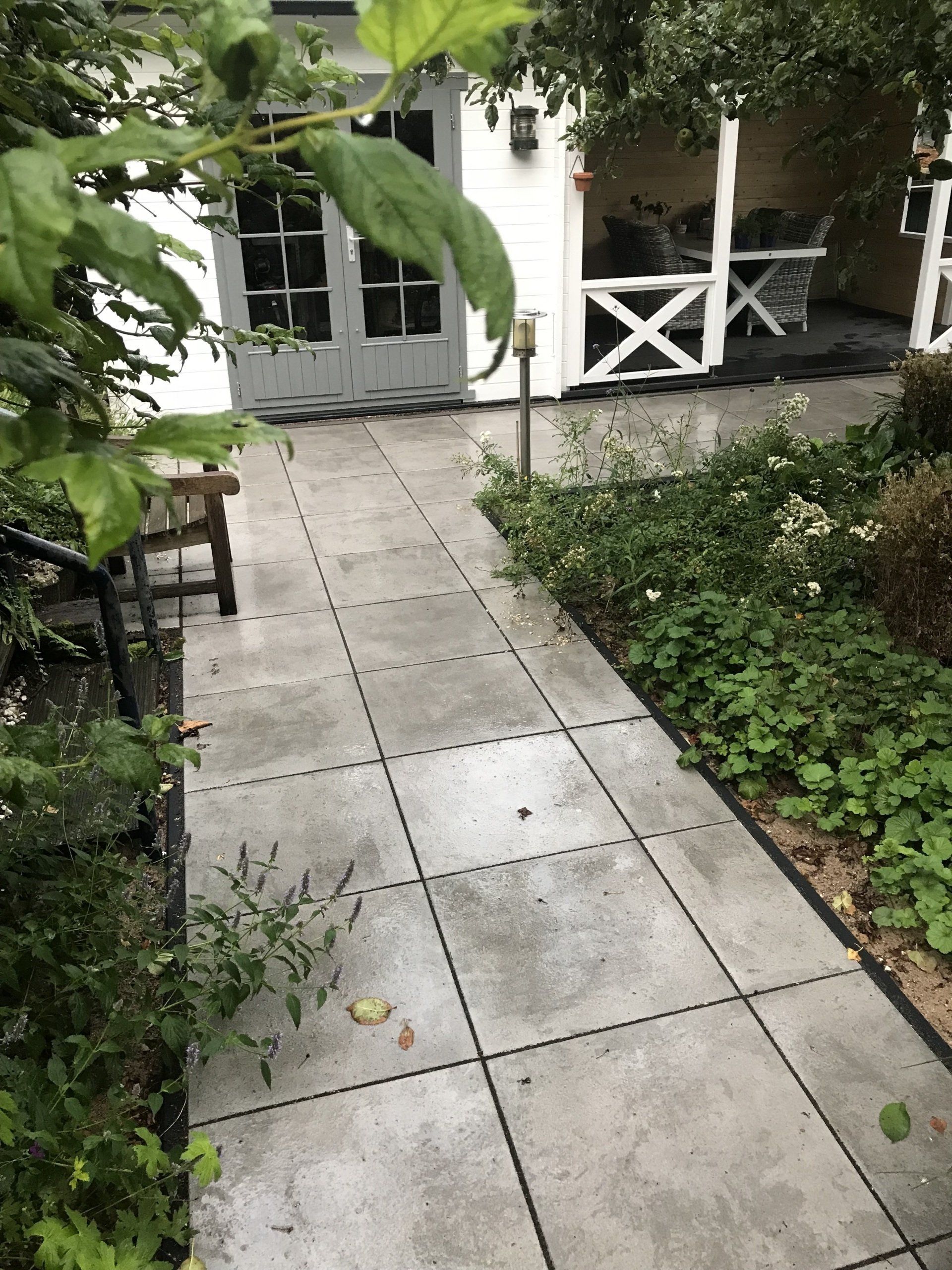 Een betonnen pad leidt naar een wit gebouw met grijze deuren, omgeven door groen en een kleine houten bank.