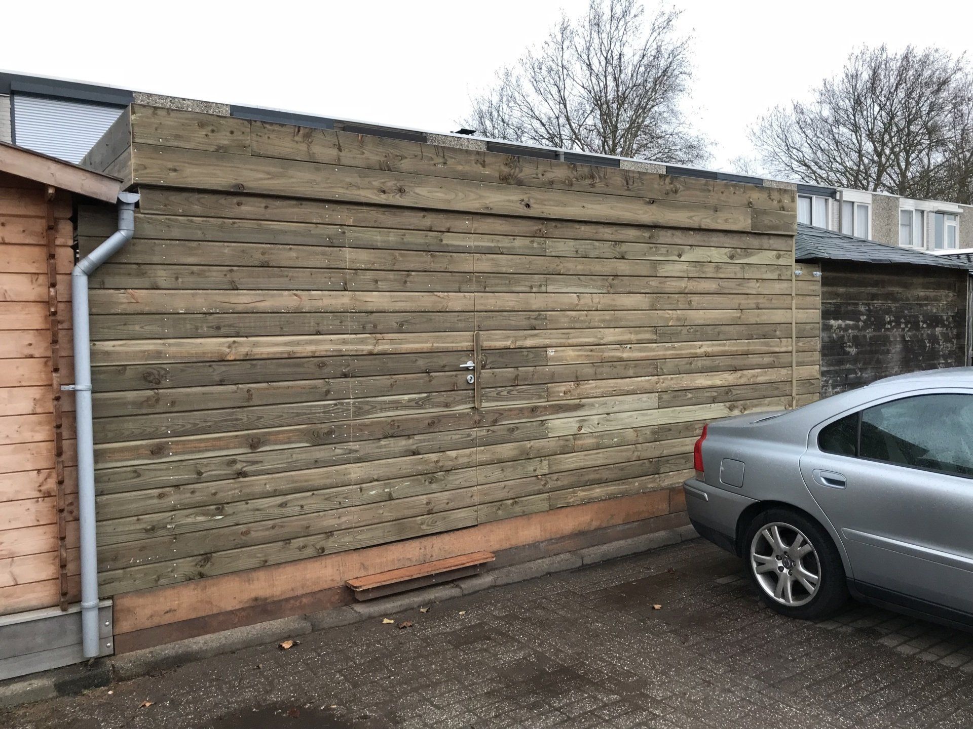 Een houten schutting naast een zilverkleurige auto en een klein schuurtje. Grijze lucht erboven.