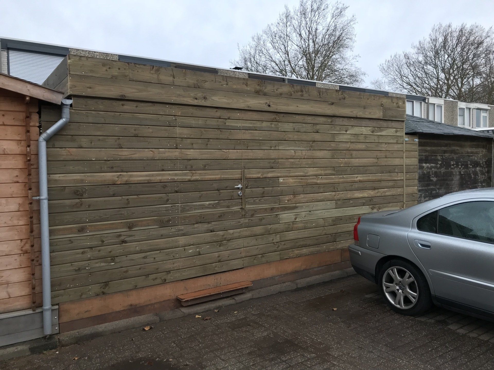 Buitenaanzicht van een muur die is geïsoleerd met horizontale, verweerde houten planken; rechts staat een auto geparkeerd.