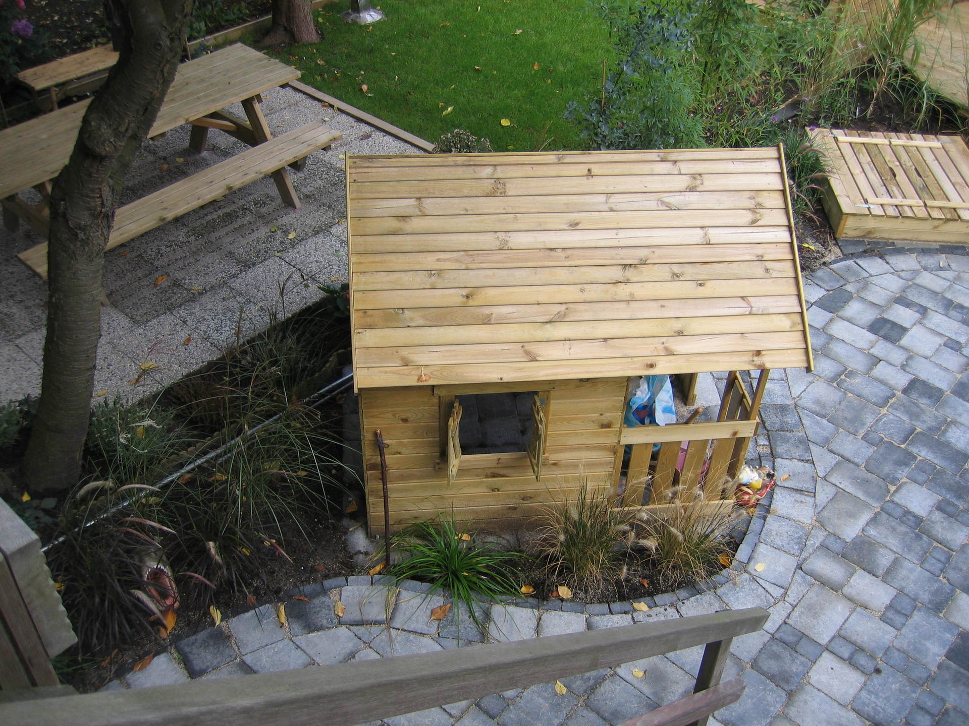 Houten speelhuisje met klein raam en schutting in de achtertuin, met een picknicktafel en bestrating.