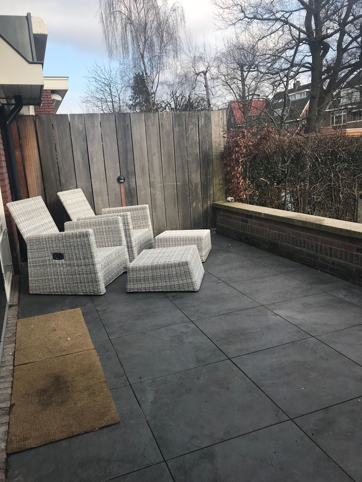 Terras met grijze rieten meubels, donkere vierkante vloertegels, houten schutting, bakstenen muur en kale bomen.