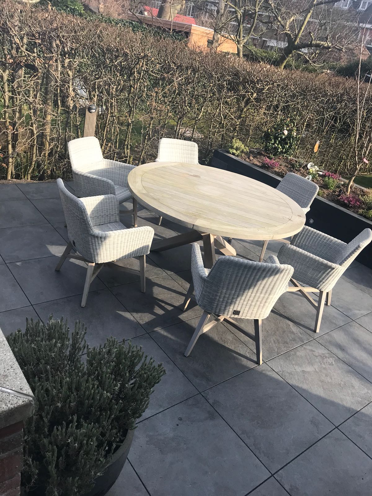 Terrasmeubilair: ovale tafel en zes witte rieten stoelen op een stenen terras met uitzicht op groen.