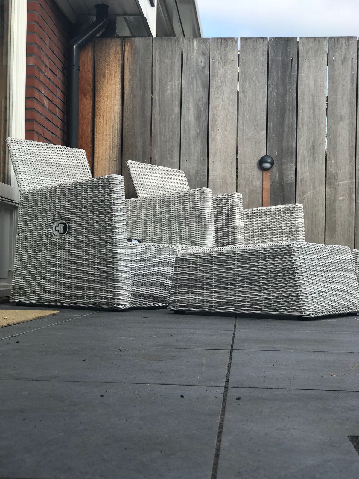 Terrasmeubilair: grijs-witte geweven hoekbank op een donkergrijze ondergrond, met een houten schutting op de achtergrond.