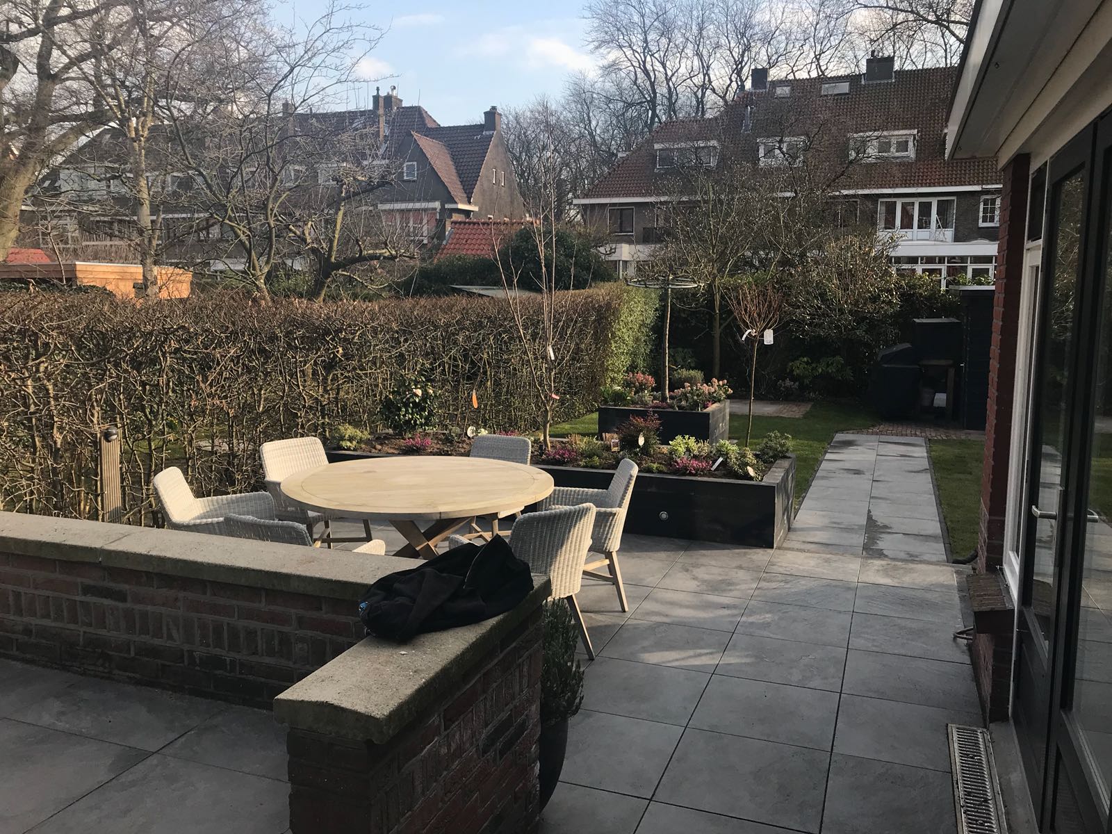 Terras met een ronde tafel, stoelen, een bakstenen muur en bloembedden; huizen op de achtergrond.