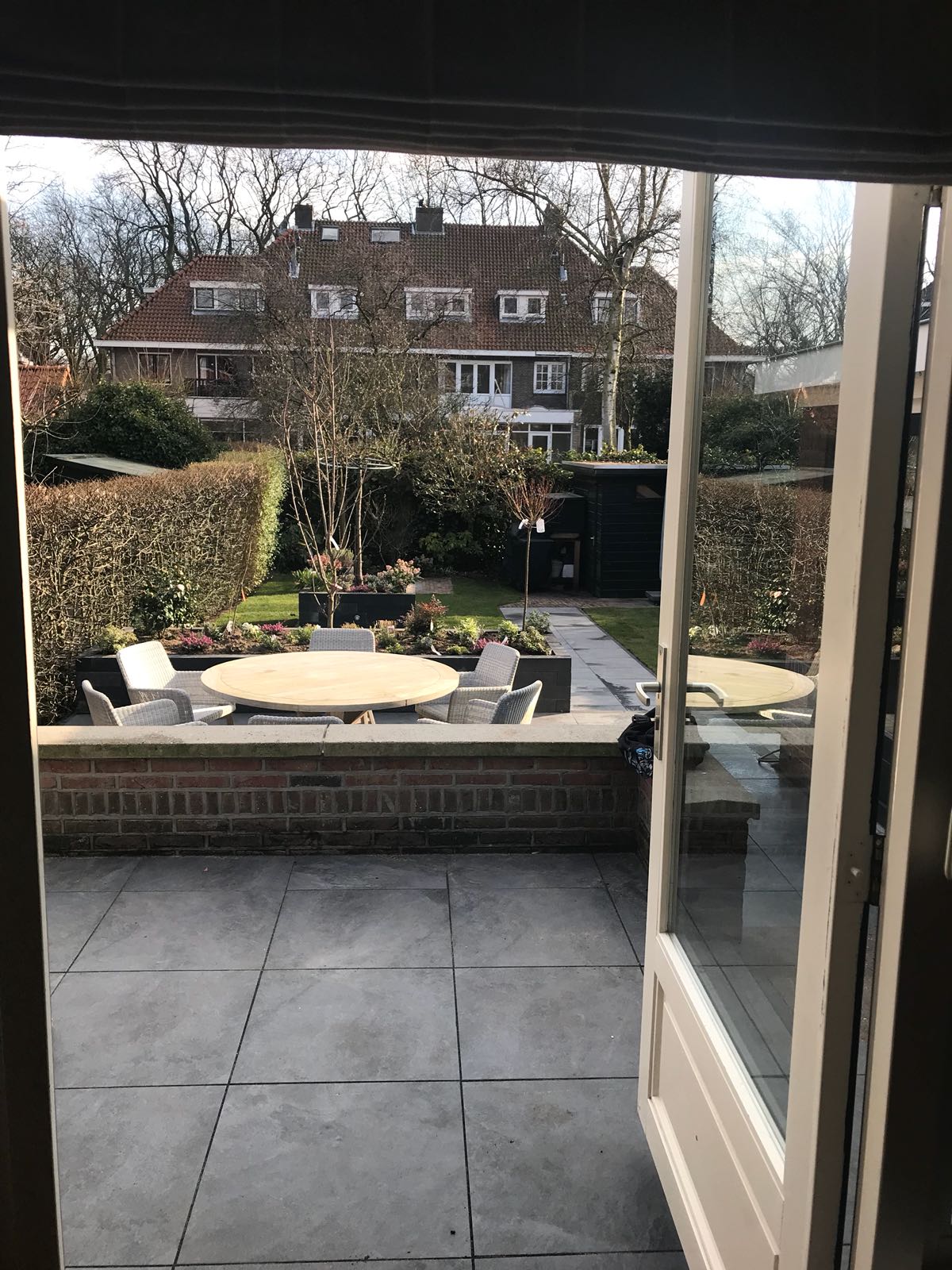 Uitzicht door openslaande deuren op een achtertuin met een terras, tafel, stoelen en een tuin, met huizen op de achtergrond.