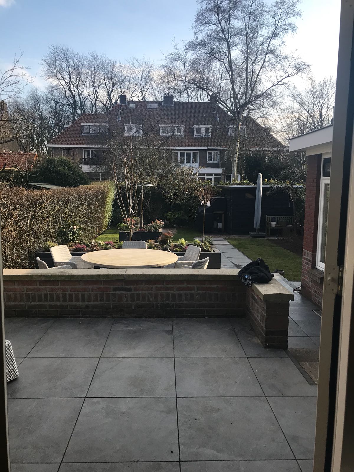 Uitzicht vanuit de achtertuin: terras met bakstenen muur, tafel en bomen. Huizen op de achtergrond onder een blauwe hemel.