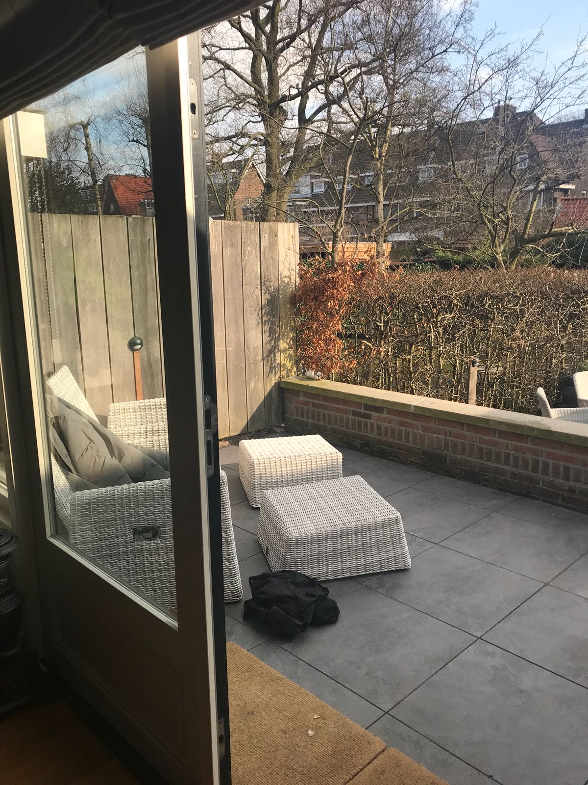 Terras met rieten meubels, bakstenen muur en groene haag, zichtbaar vanuit binnen.