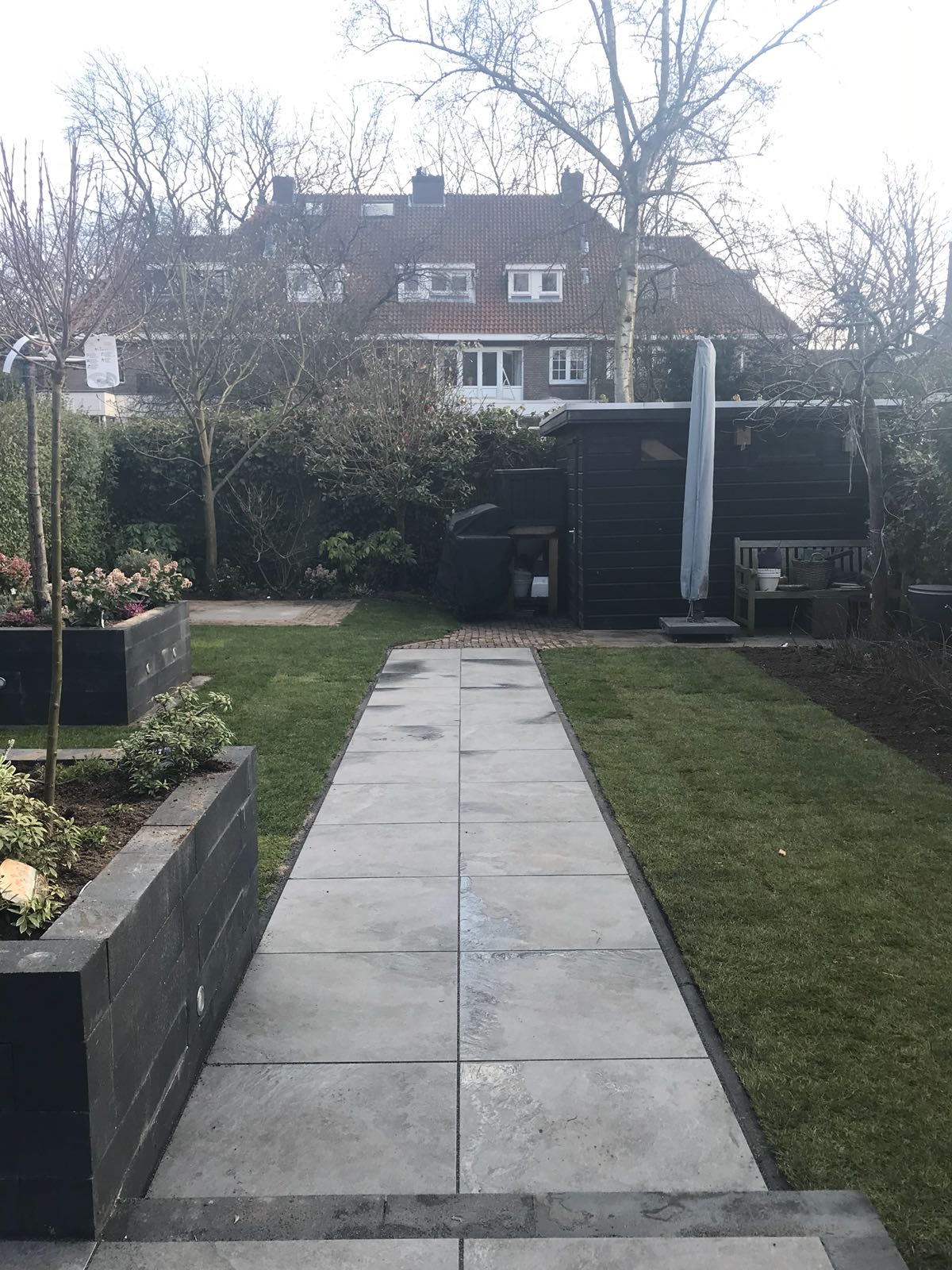 Een stenen pad loopt door een tuin met verhoogde bloembedden, een gazon en een donker schuurtje. Op de achtergrond is een huis te zien.