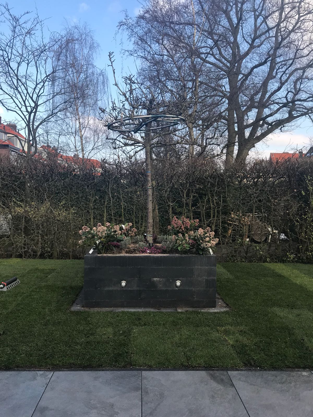 Monument met een metalen kruis en bloemen, geplaatst tegen een heg en bomen, op een grasveld.