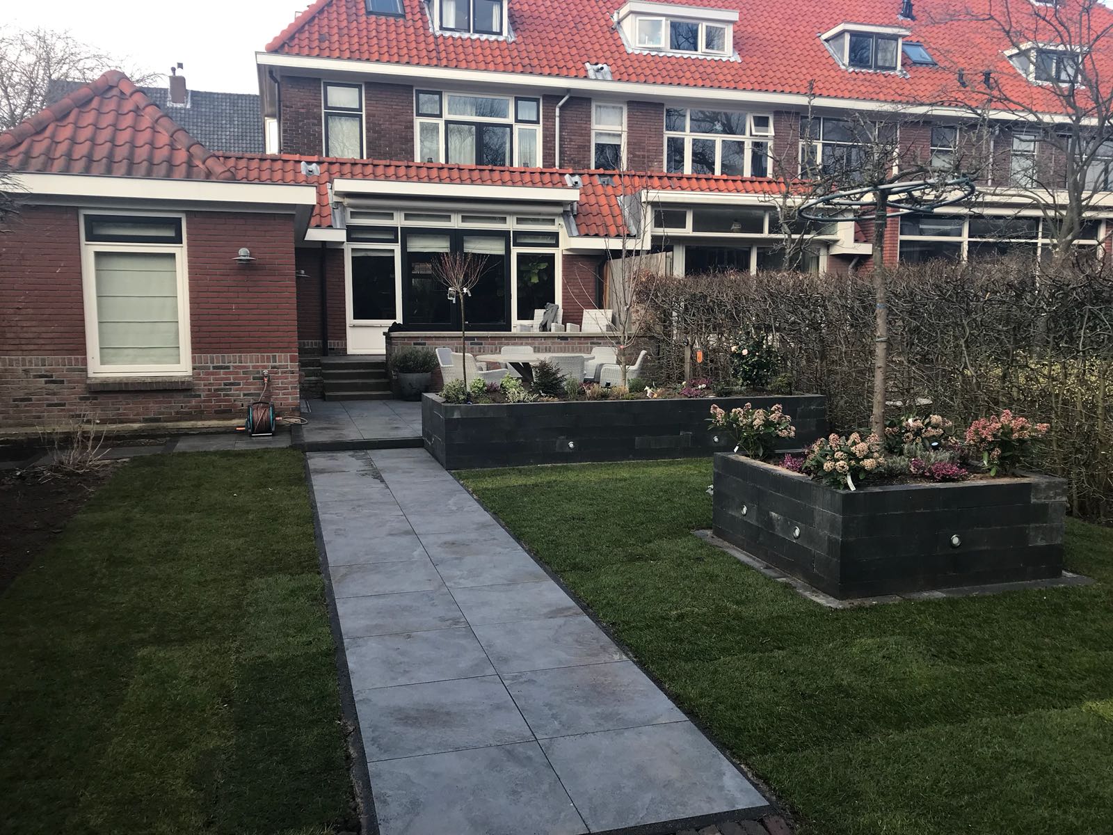 Een bakstenen gebouw met een rood pannendak, en een grijs pad leidt naar het huis, met een gazon en plantenbakken.