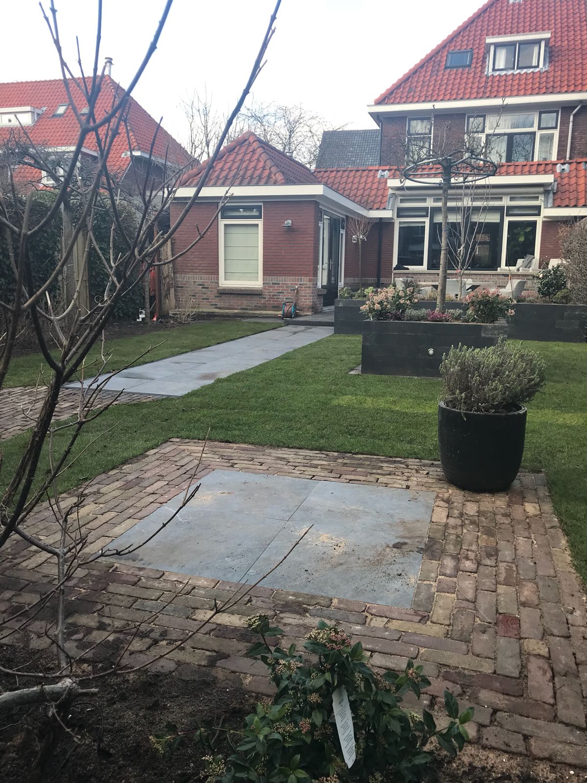 Achtertuin met stenen terras, gazon en pad naar stenen gebouw. ​​Huizen met rode daken op de achtergrond. Bewolkte lucht.