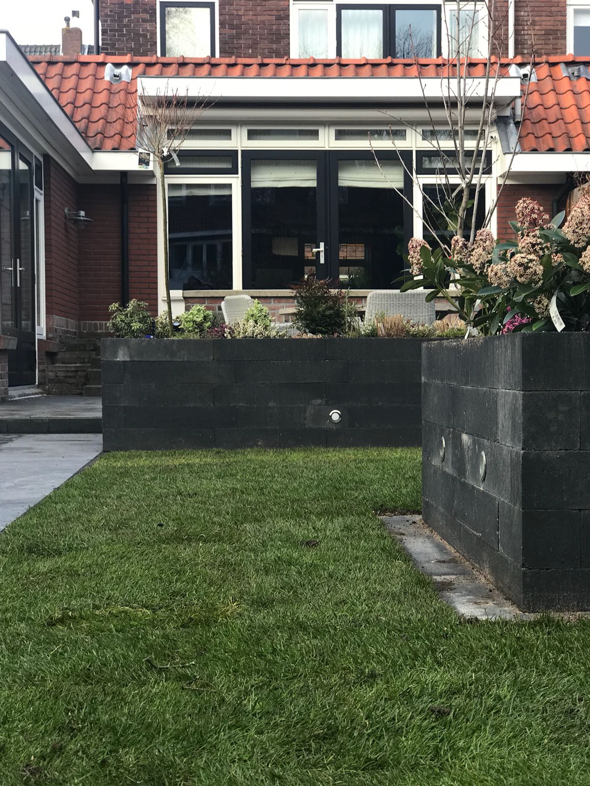 Een voortuin met een rechthoekige, donkergrijze plantenbak, een gazon en een gebouw met een rood pannendak.
