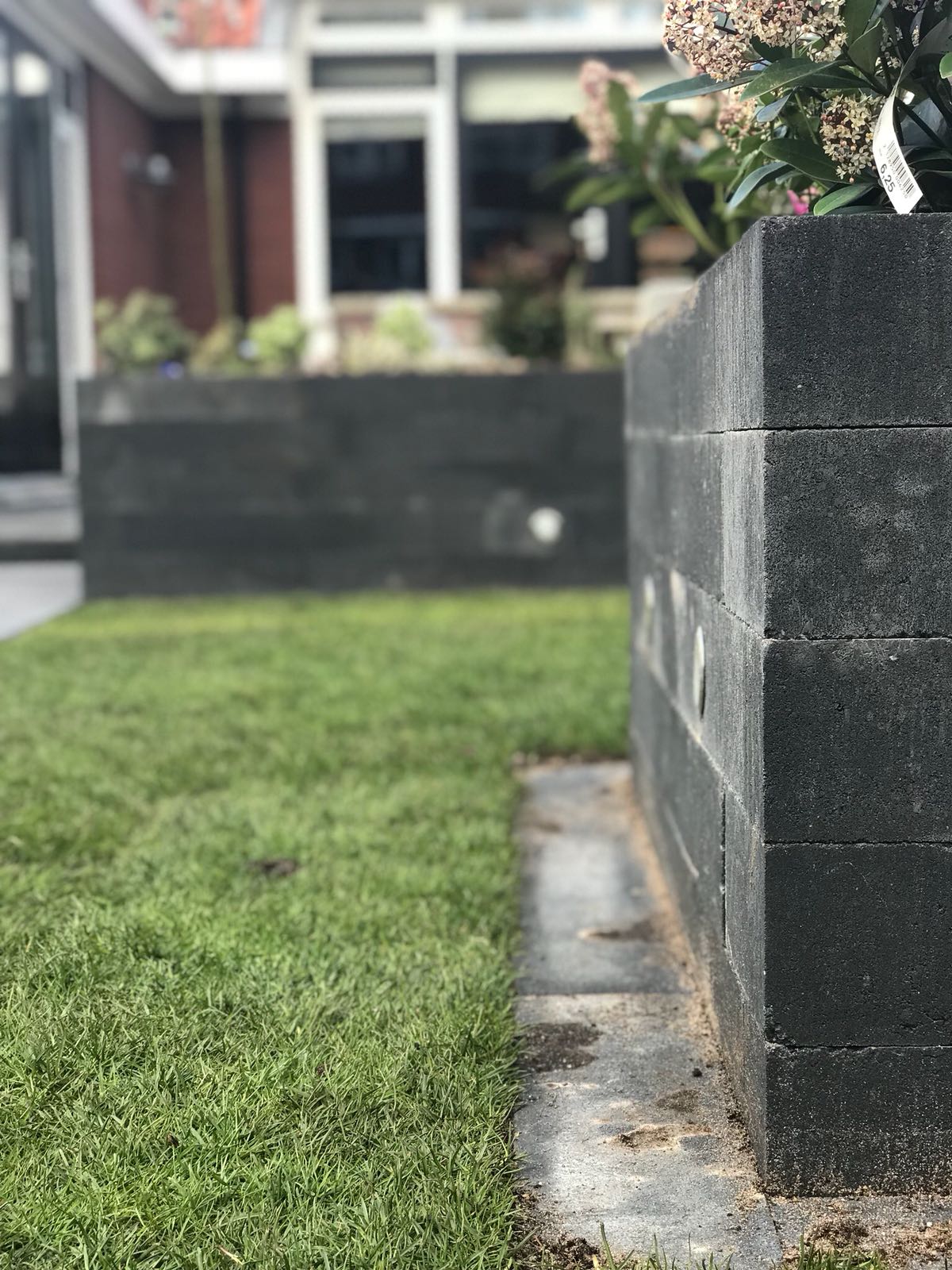 Donkergrijze bakstenen keermuren omringen een groen gazon; op de achtergrond is een gebouw te zien.
