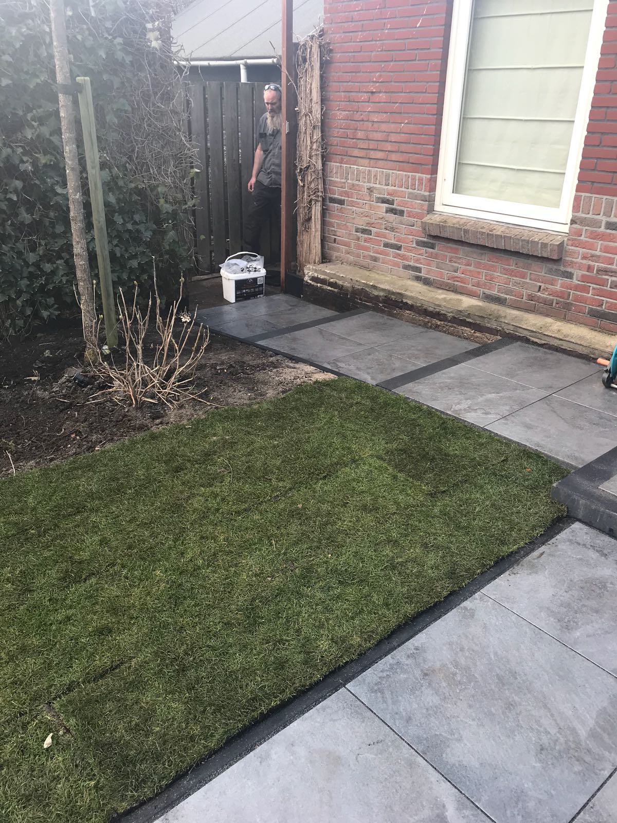 Achtertuin met donkere bestrating en gras. Persoon bij het hek, bakstenen muur en raam aan de rechterkant.