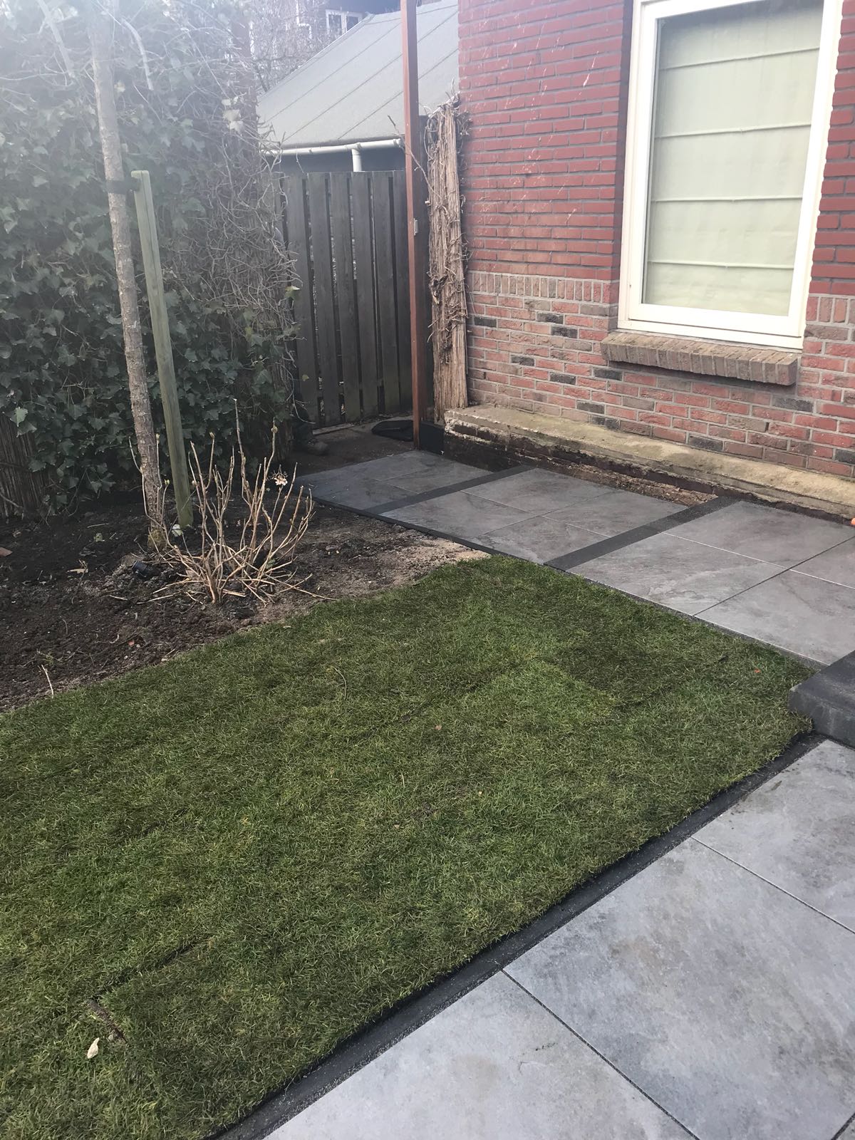Een groen grasveldje met een stenen pad, een houten poort en een bakstenen gebouw.