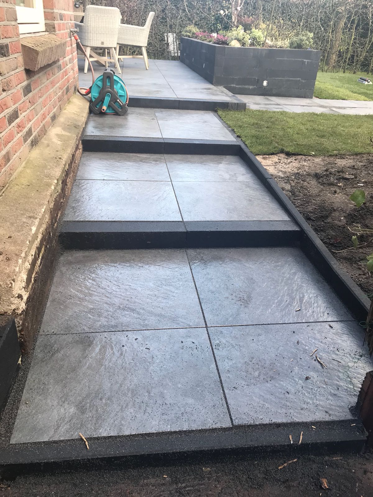 Een betonnen buitentrap leidt naar een terras, omzoomd door donker hout en pas aangelegd gras.