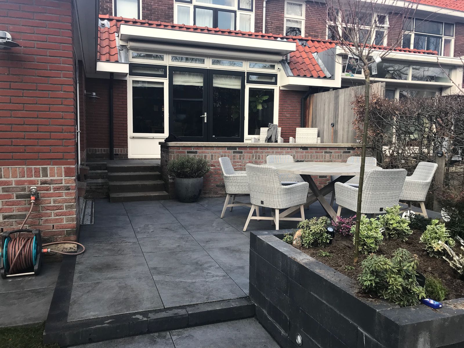 Terras met tuinmeubilair en bloemperk, dat via een donkere deur toegang geeft tot het huis.