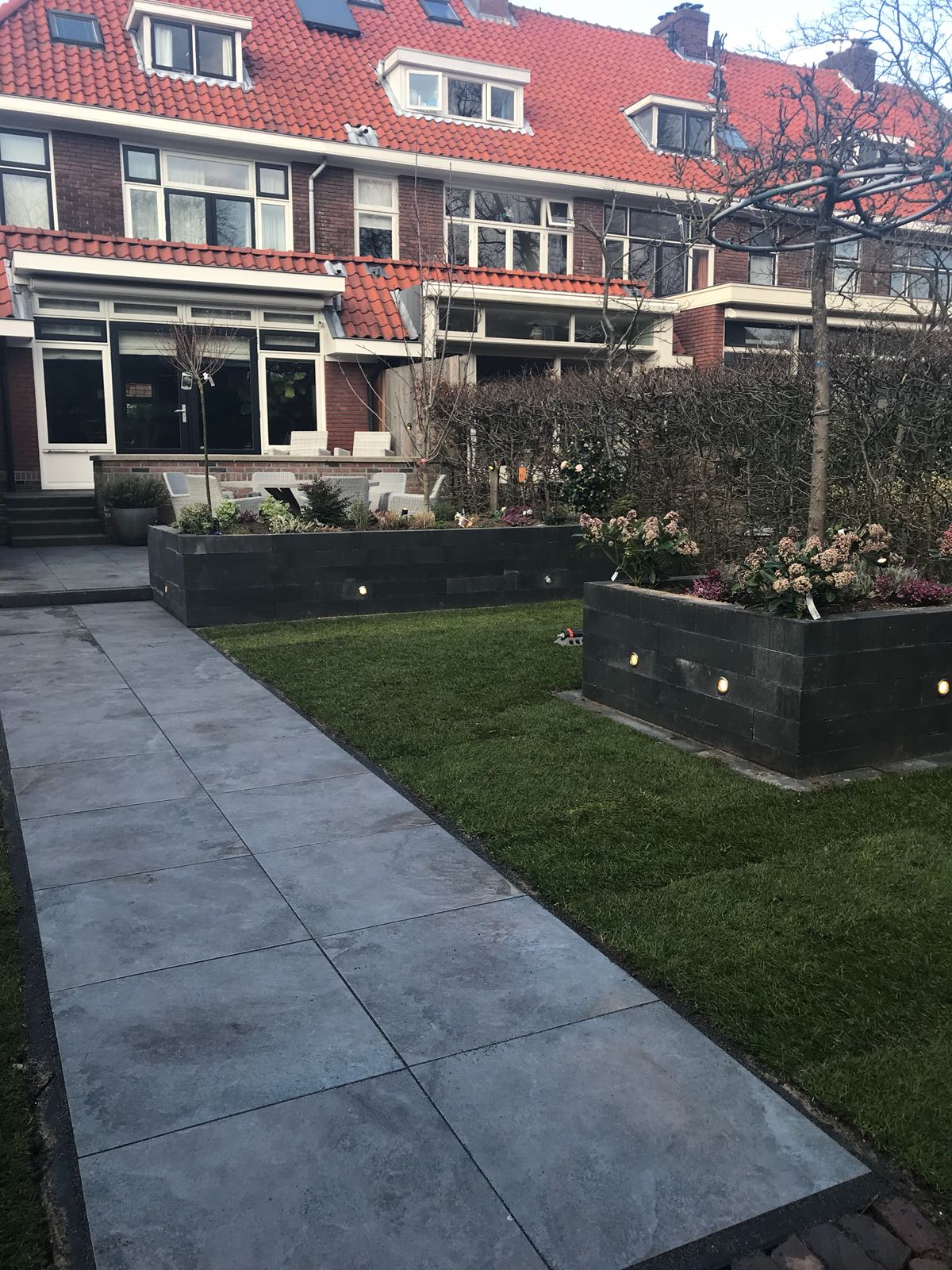 Een grijs stenen pad leidt naar een bakstenen huis met een tuin. Langs het pad staan ​​donkere plantenbakken met bloeiende struiken.