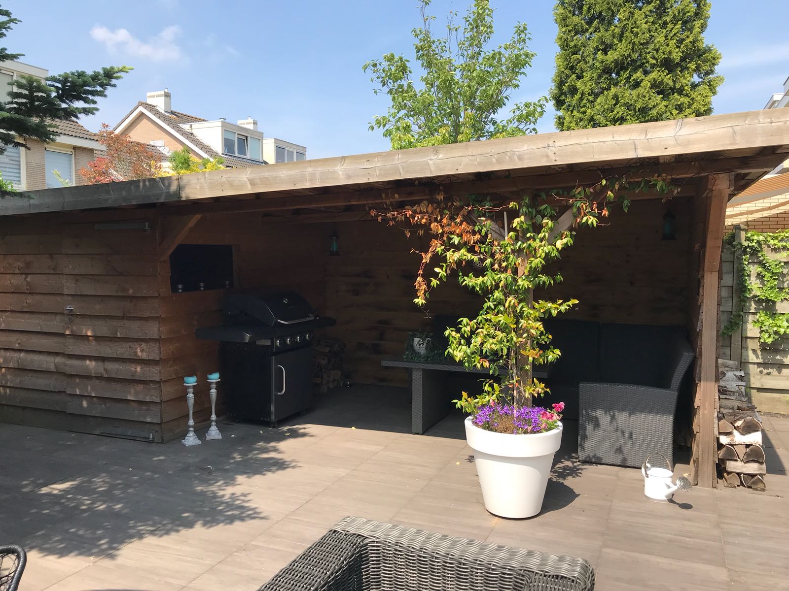 Terras met houten afdak, barbecue, zitgelegenheid, potplant en een witte pot met bloemen. Zonnige dag.