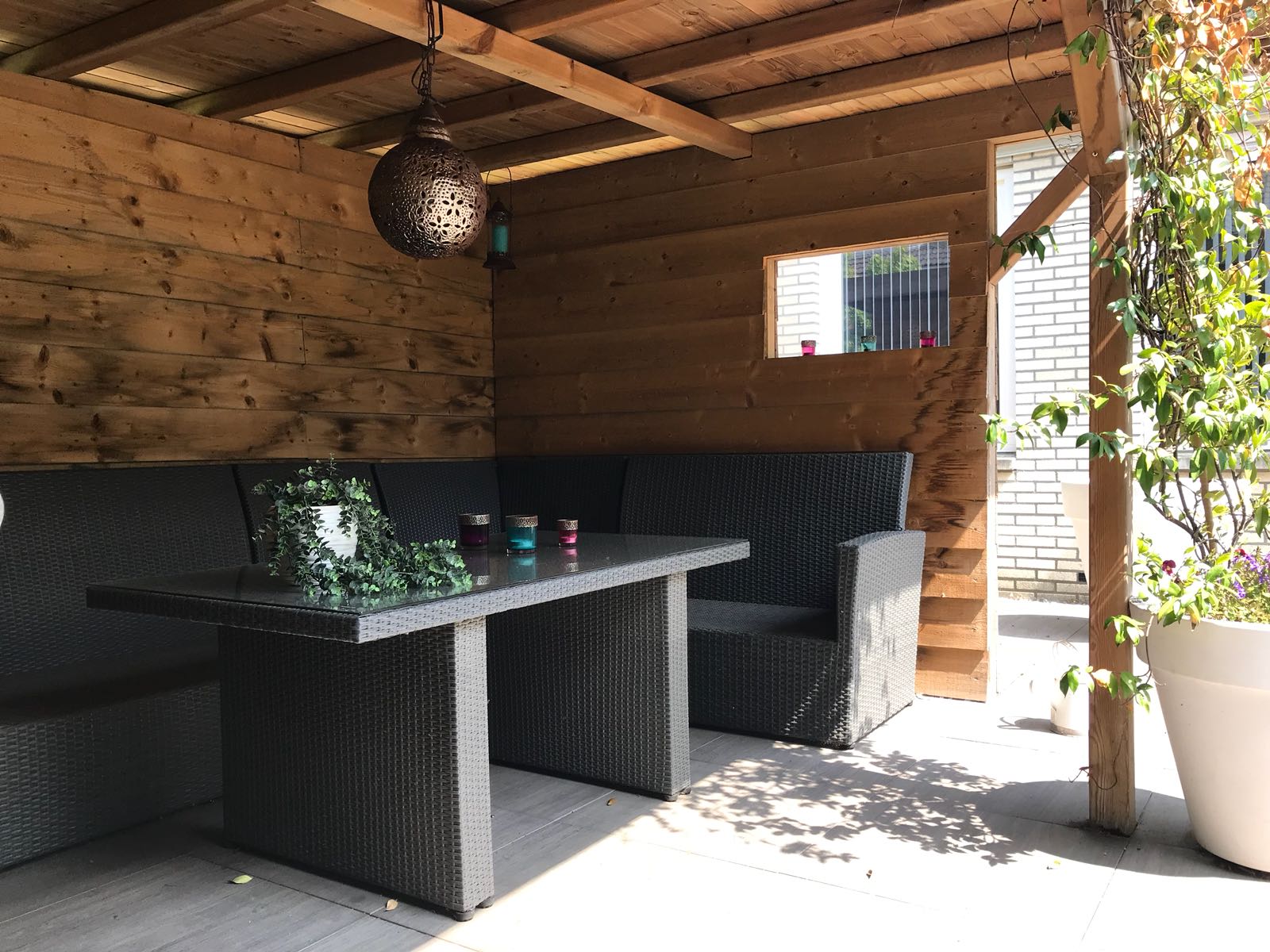Overdekt terras met tafel en stoelen; houten lambrisering, sfeerverlichting, groen en potplanten.