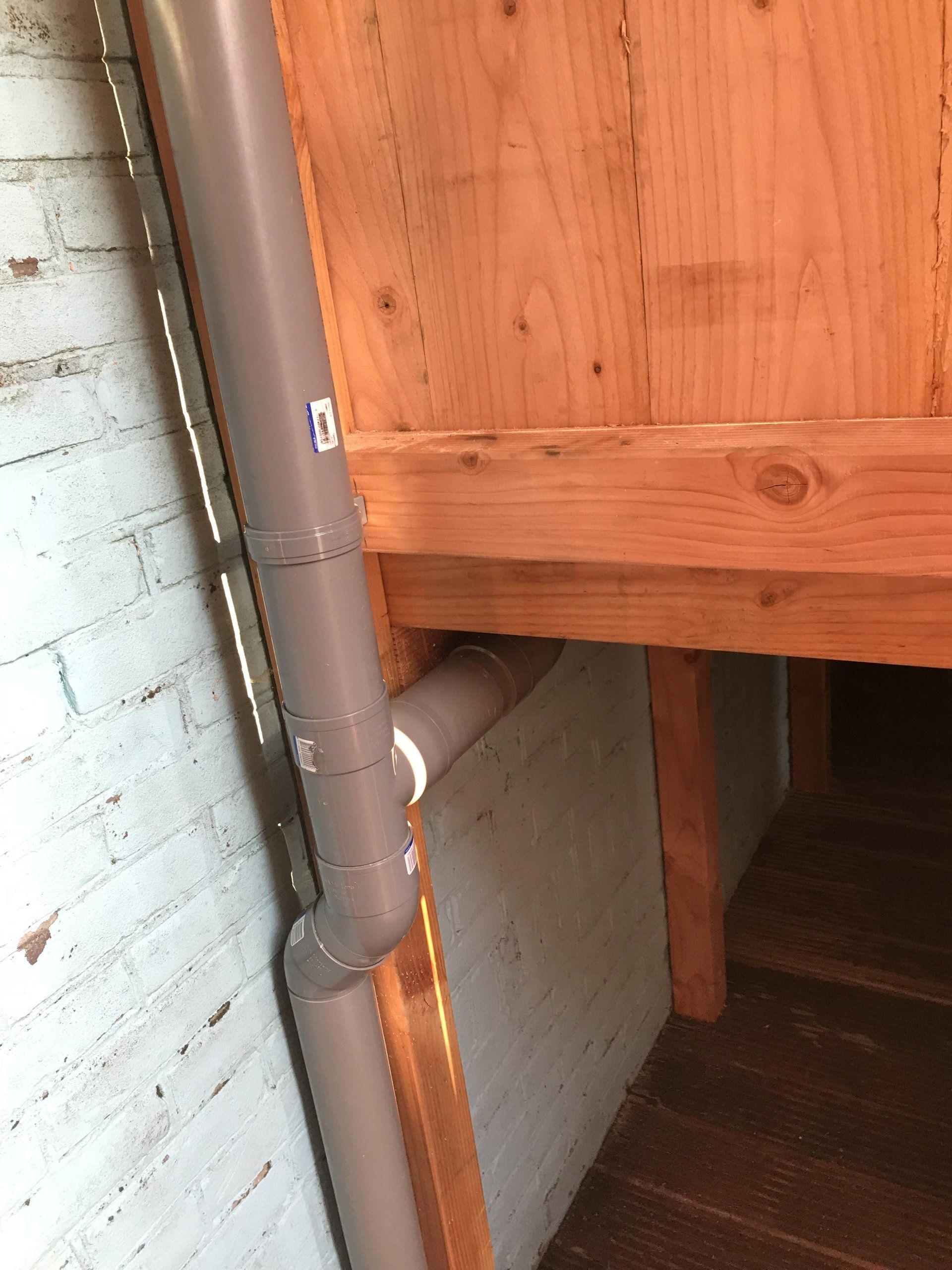 Grijze PVC-buizen die aansluiten op een houten constructie, tegen een witte bakstenen muur.