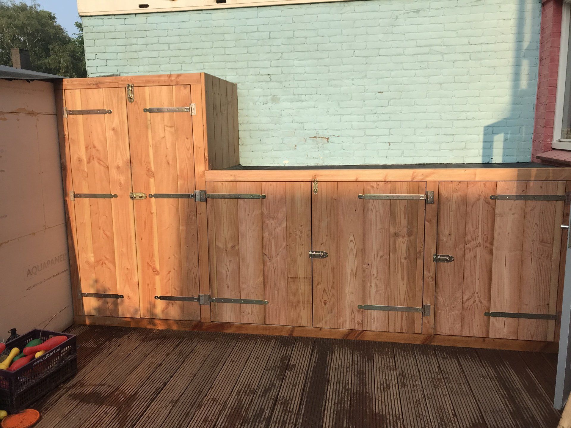 Houten buitenopbergkast tegen een lichtblauwe bakstenen muur.