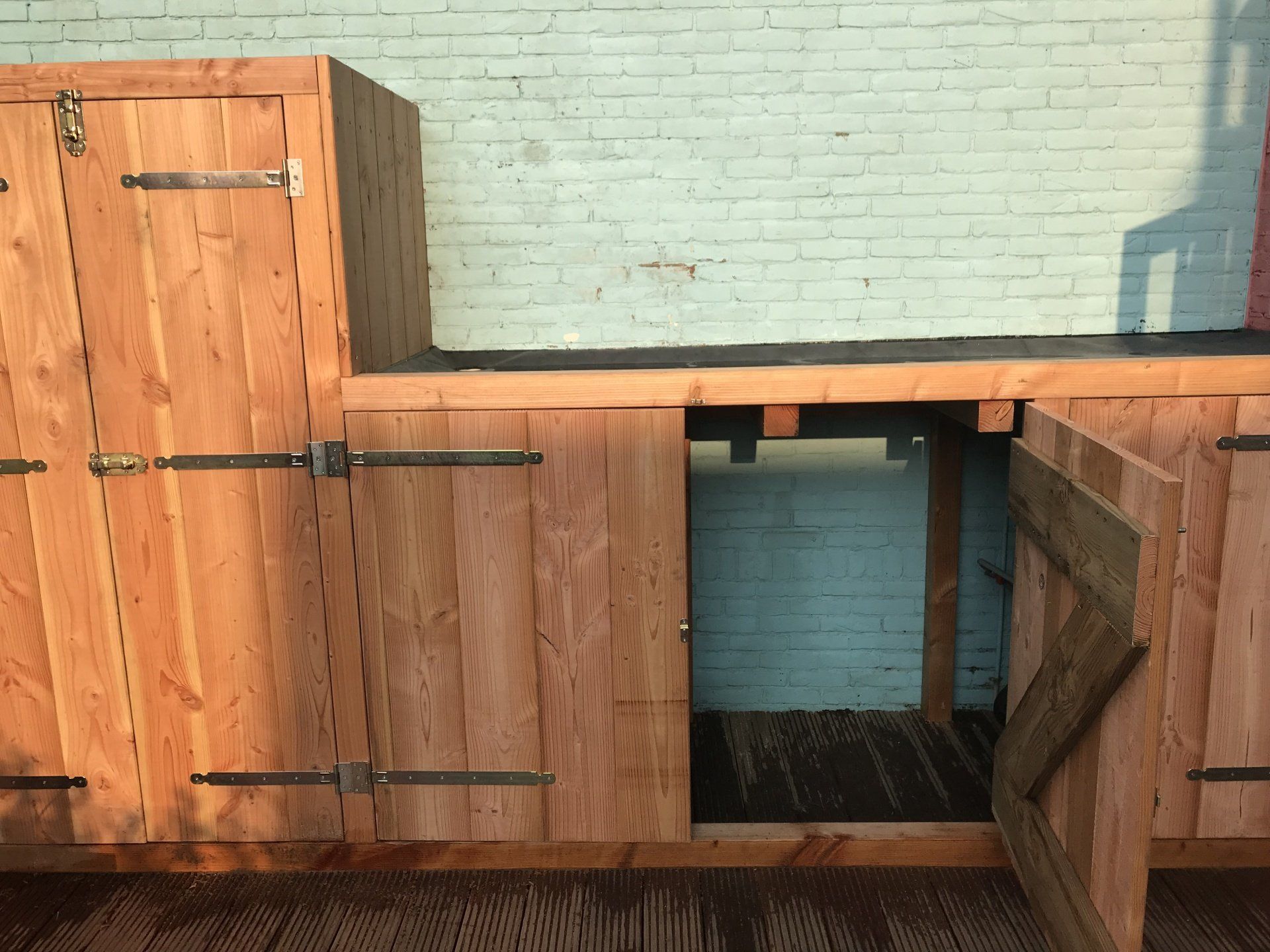 Houten buitenopbergkast met openslaande deuren tegen een lichtblauwe bakstenen muur.