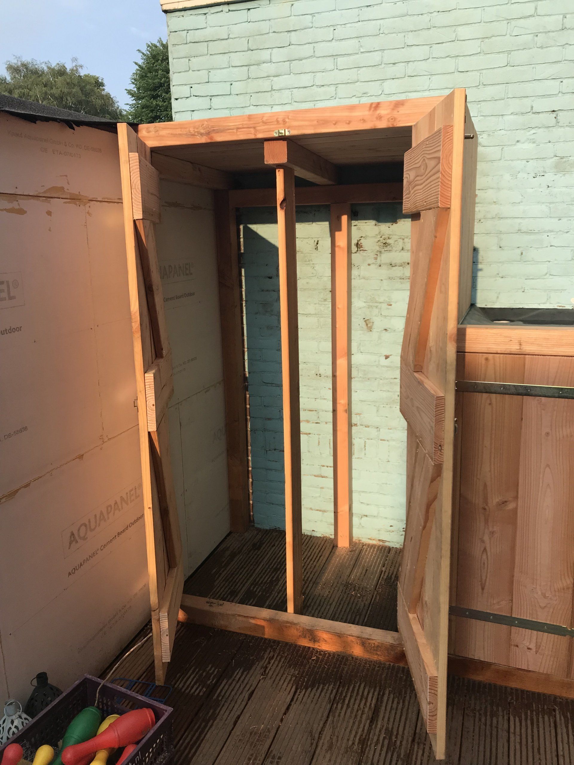 Houten frame voor een buitenopbergunit met een lichtblauwe gebouwachtergrond.