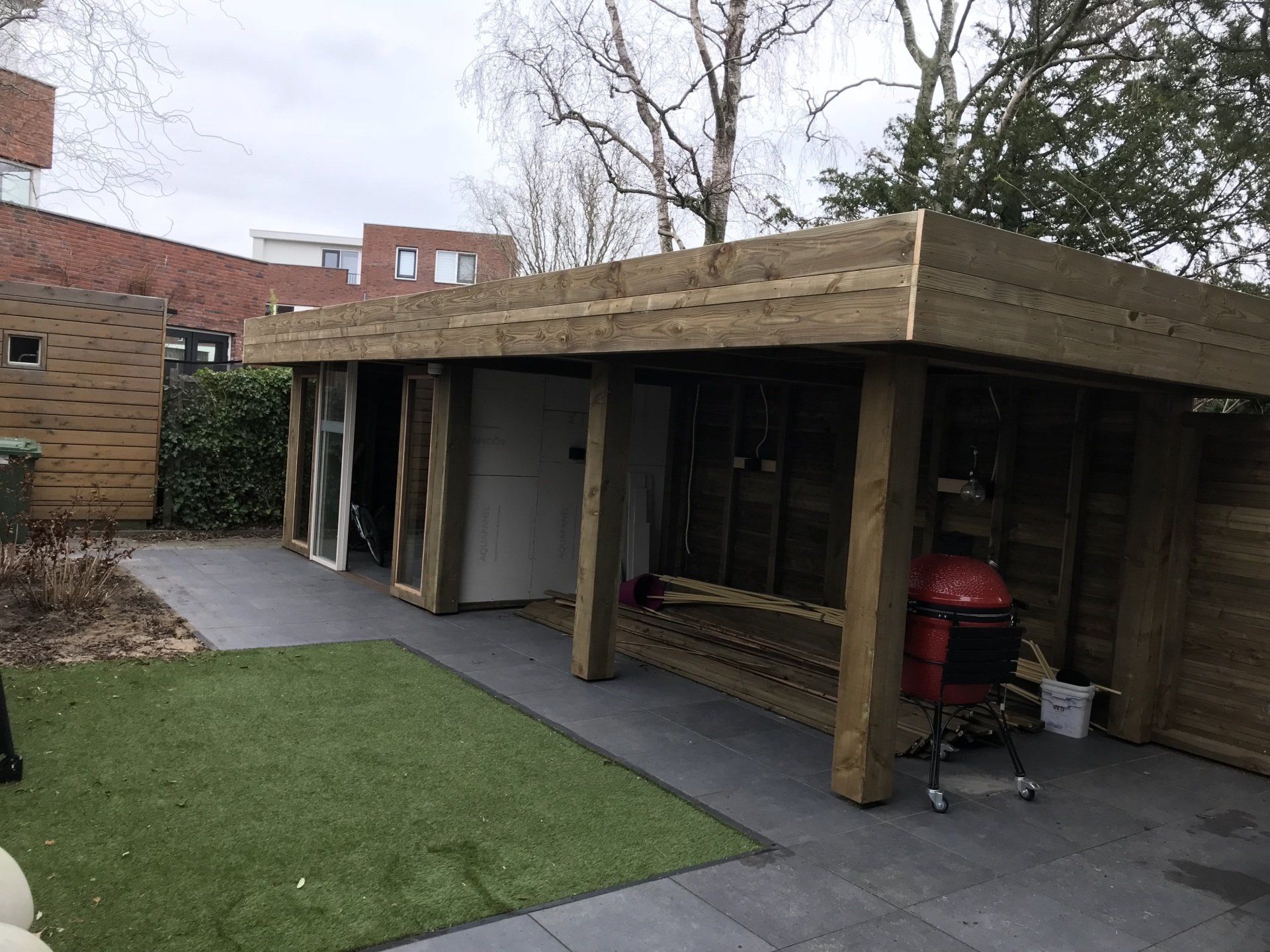Houten schuur met open zijkanten, gebouwd op een terras met een rode barbecue zichtbaar. Op de achtergrond zijn een groen gazon en een gebouw te zien.