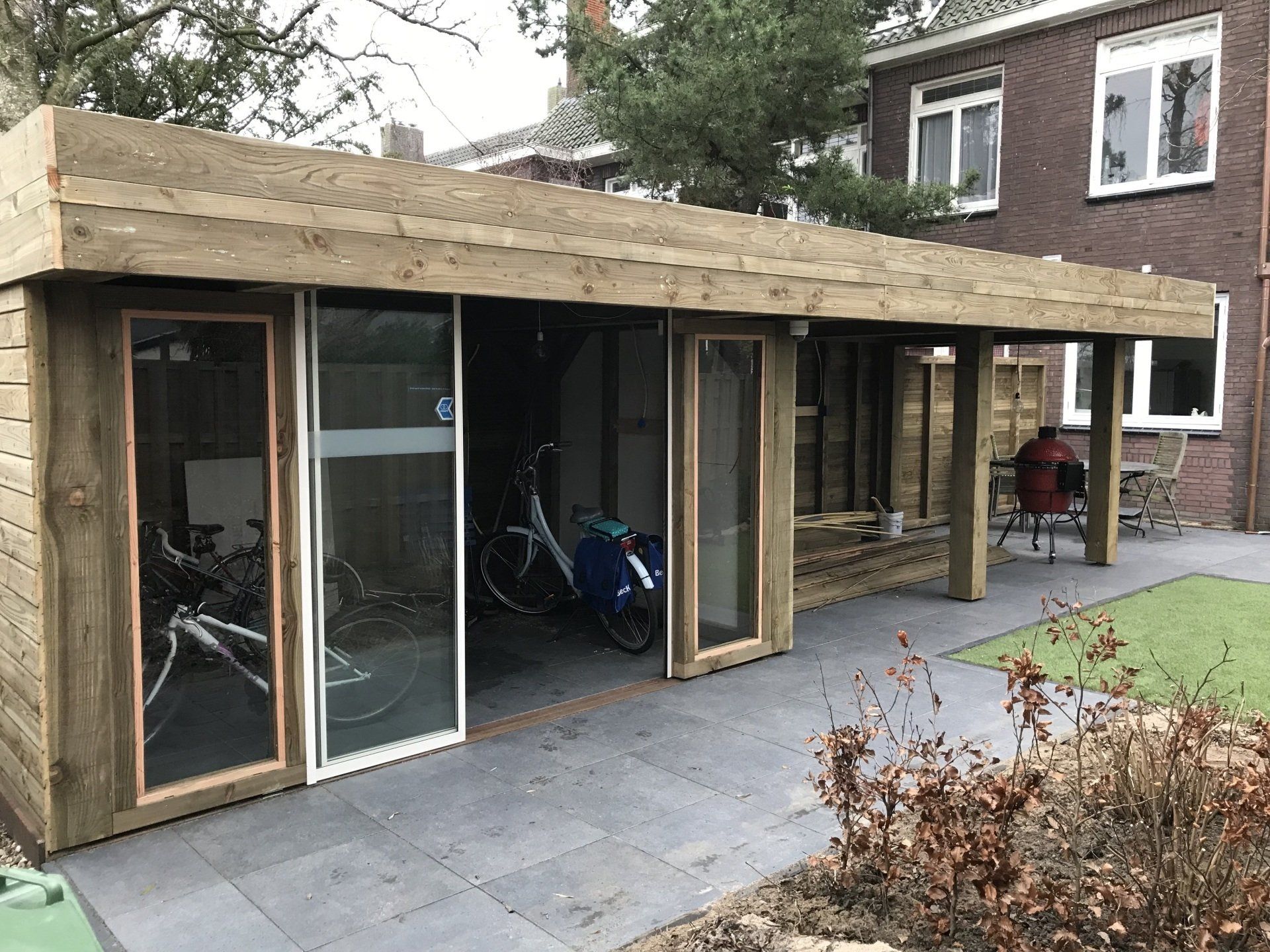 Houten schuur met glazen schuifdeuren, waarin fietsen en een zitgedeelte buiten zijn ondergebracht.