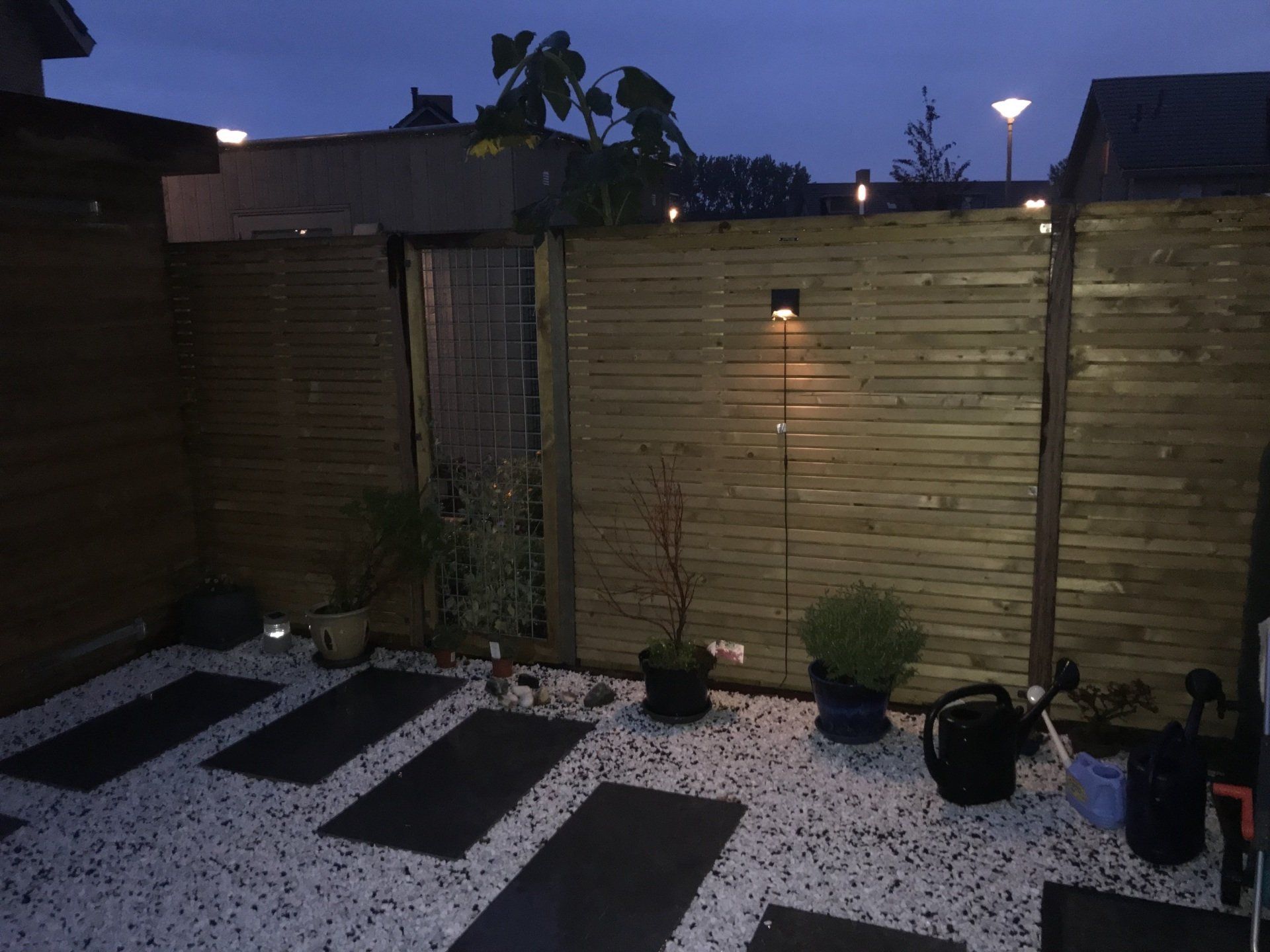 Buitenterras met houten schutting, donkere bestrating, wit grind en kleine potplanten; verlicht met sfeerverlichting.